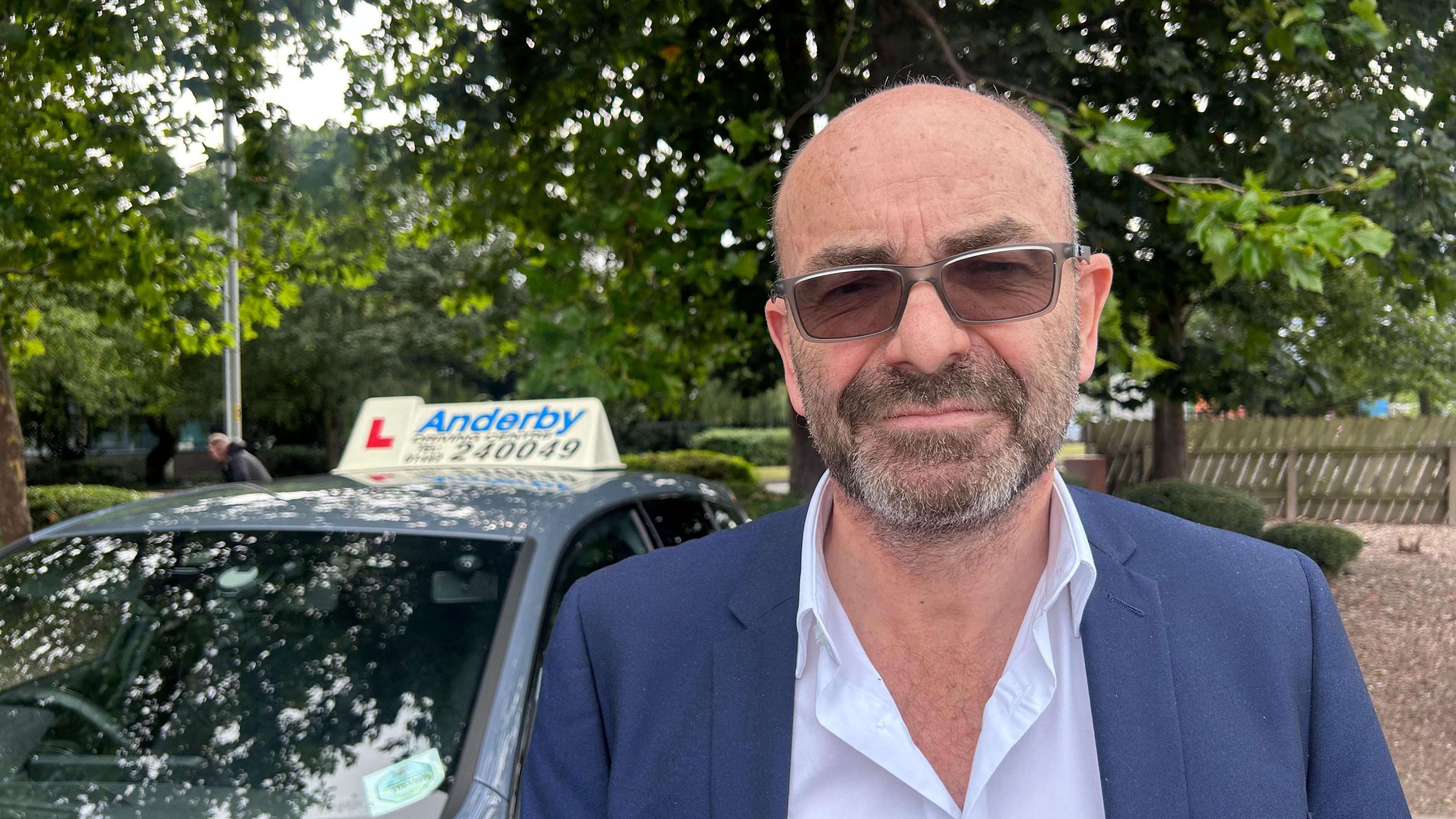 A man with tinted glasses on stood next to a blue BMW with an L plate and an advertisement board for his driving school. He has a black haired stubble with white patches and is wearing a blue blazer. The white shirt he has on has his top button un-done and he has a glum face looking at the camera. The man is stood next to his parked car in a car park.