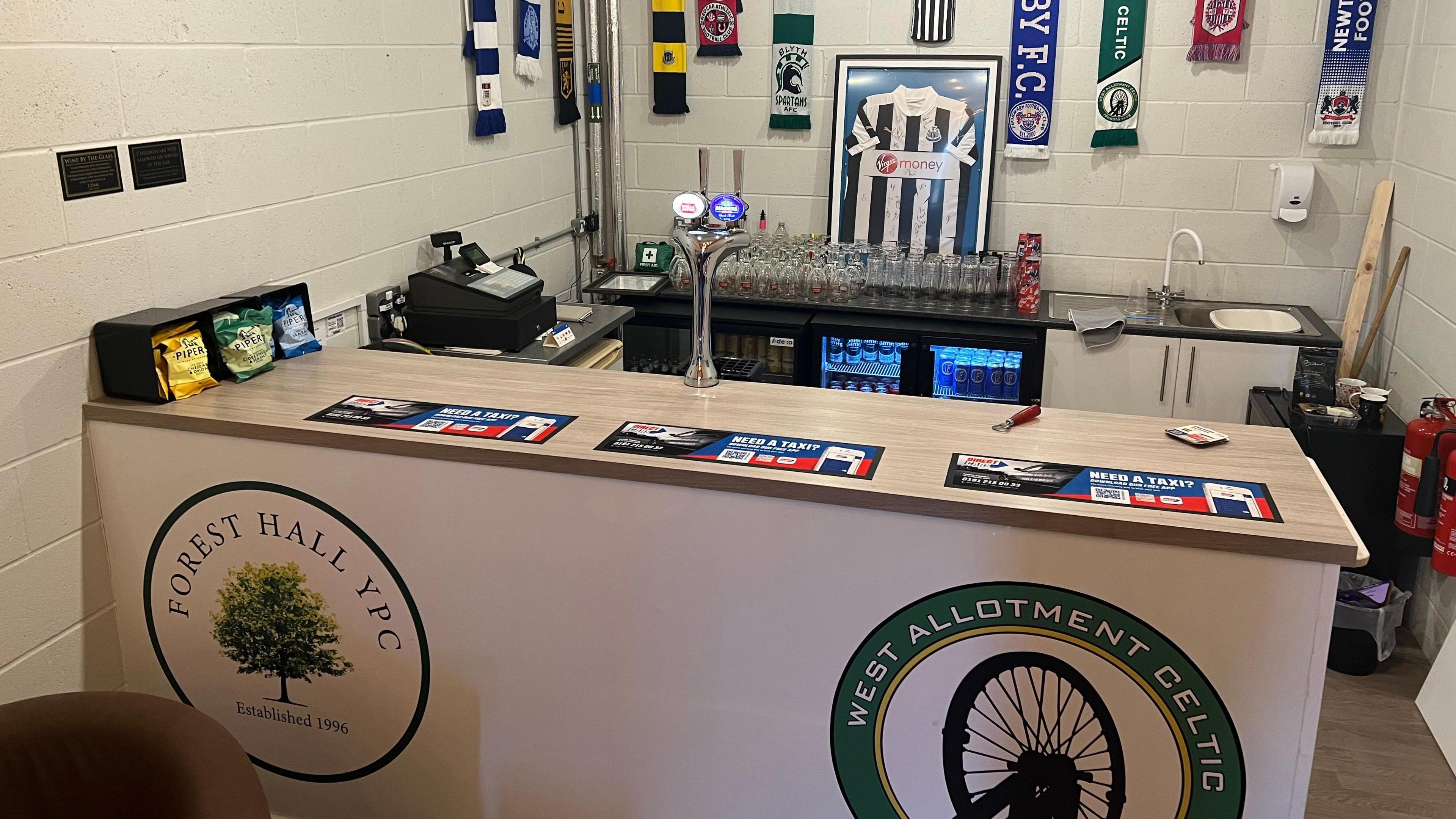 A small room with a bar including two pumps, a till and two fridges full of drinks. There is a tap to one side and football scarves hanging off the wall.