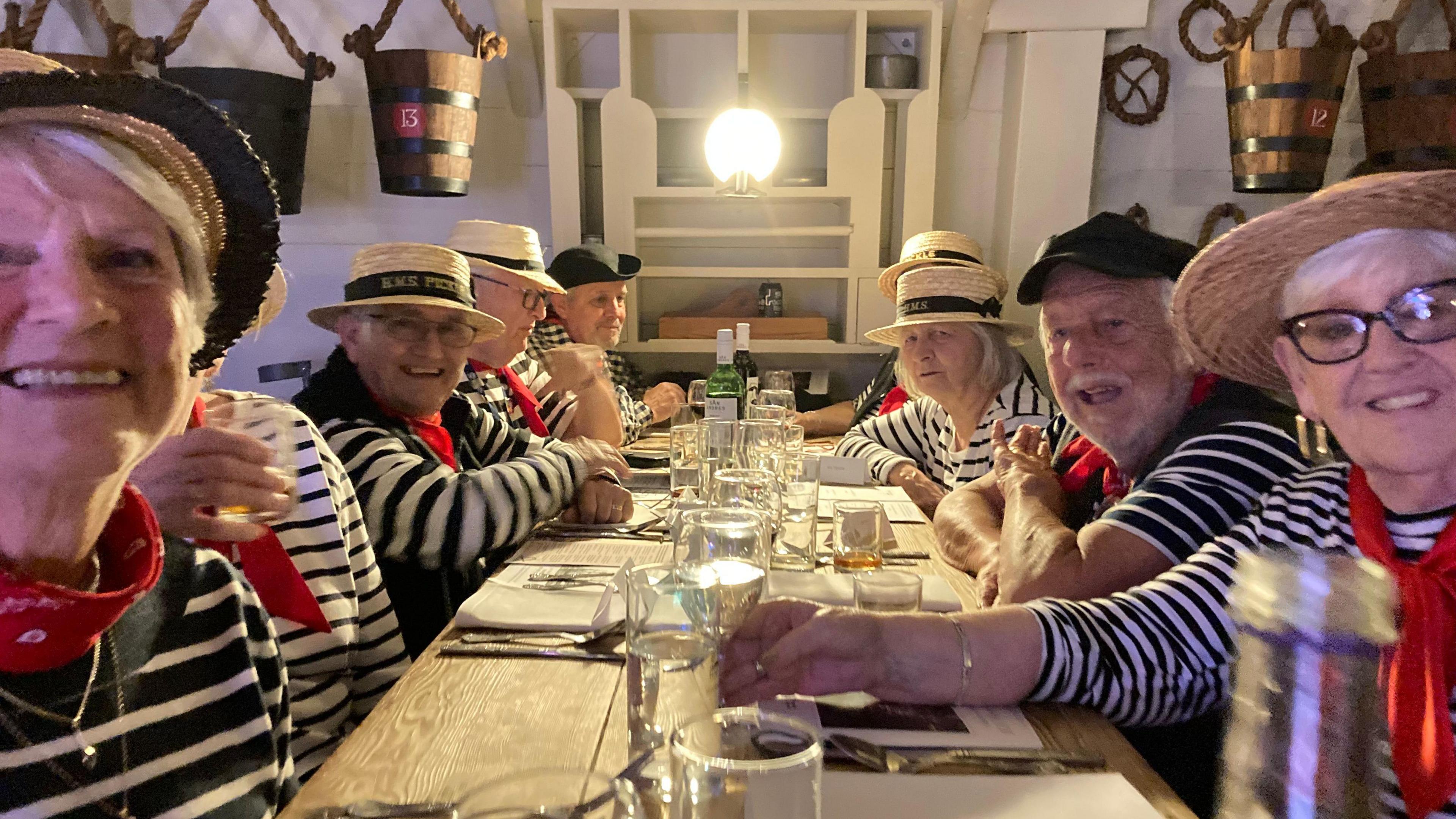 Half a dozen men and women, dressed in blue and white striped tops, red scarves, white trousers and wearing a hat, all sat around a long bench-style table, smiling at the camera