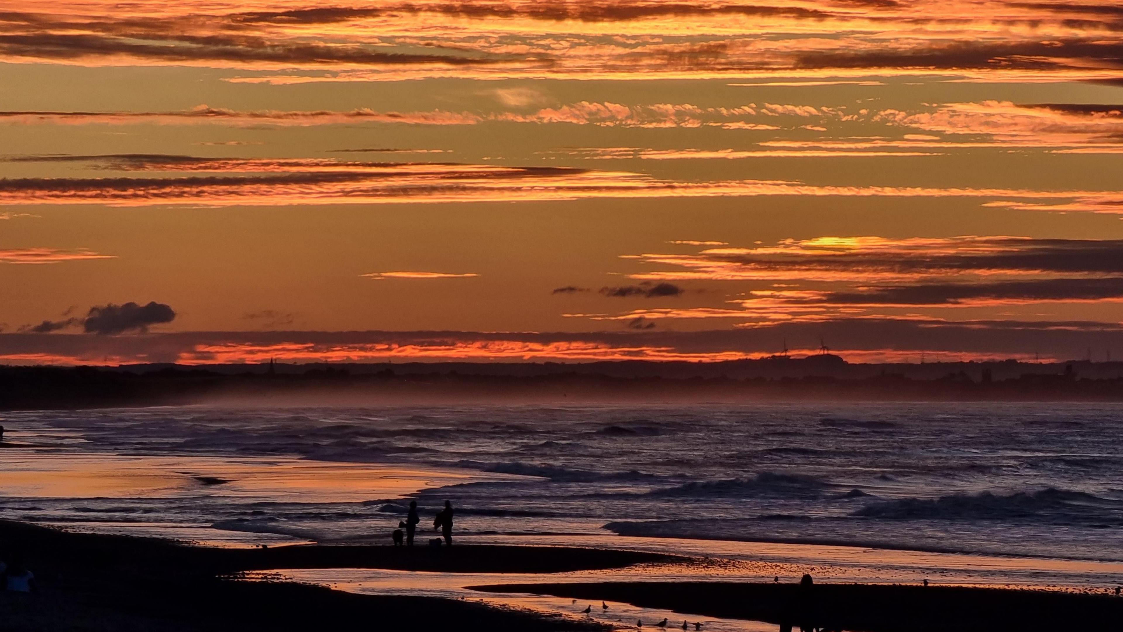 Small waves crashing onto a beach where groups of people are walking along in the distance.  The sky is filled with thin layers of cloud that have been turned orange and yellow by the sunset.