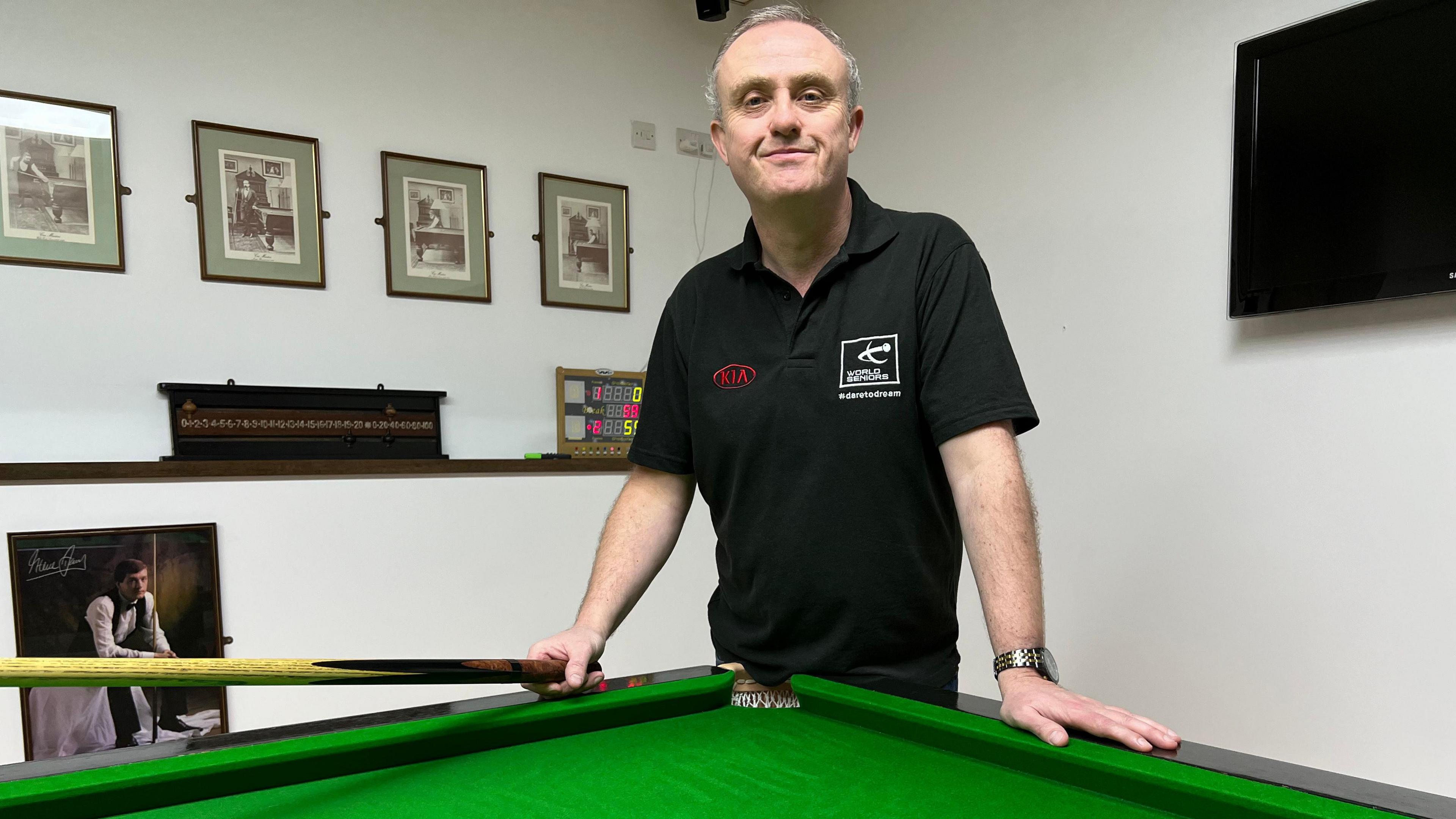 Aaron Canavan, a man with grey hair, wearing a black polo shirt with red and white badges on the chest. He is holding a snooker cue and leaning over a green snooker table, while staring directly at the camera. Behind him are black and white, framed photos arrayed on the wall. There is black television on the wall over his left shoulder.