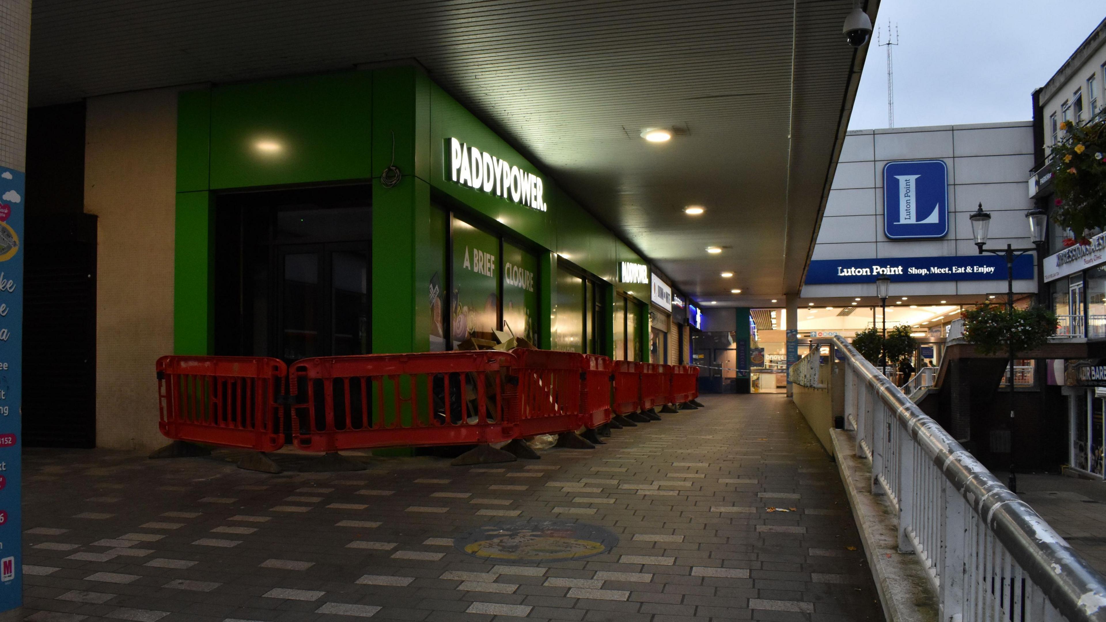 An outdoor ramp leading up to Luton Point shopping centre. There are grey and white bricks on the floor and a metal railing on the right. There is a row of shops on the left of the ramp, the main one in shot is Paddy Power with a green exterior and white sign. It is quite gloomy with a grey sky.