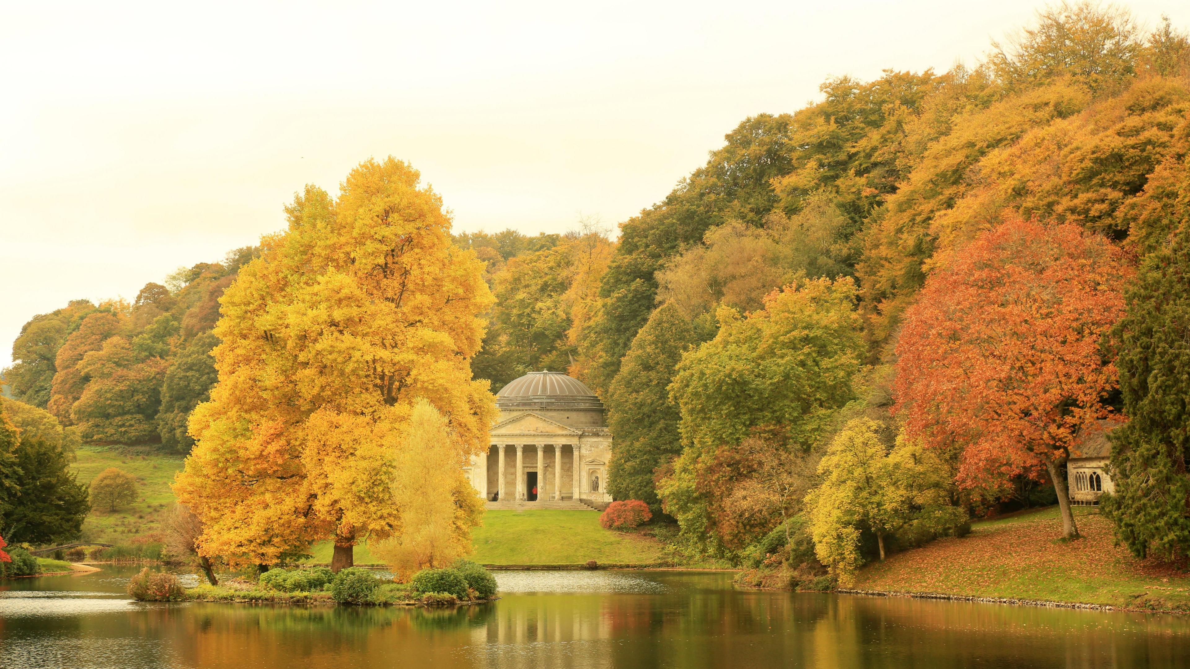 A large lake surrounded by orange and green trees and an old pantheon with pillars.
