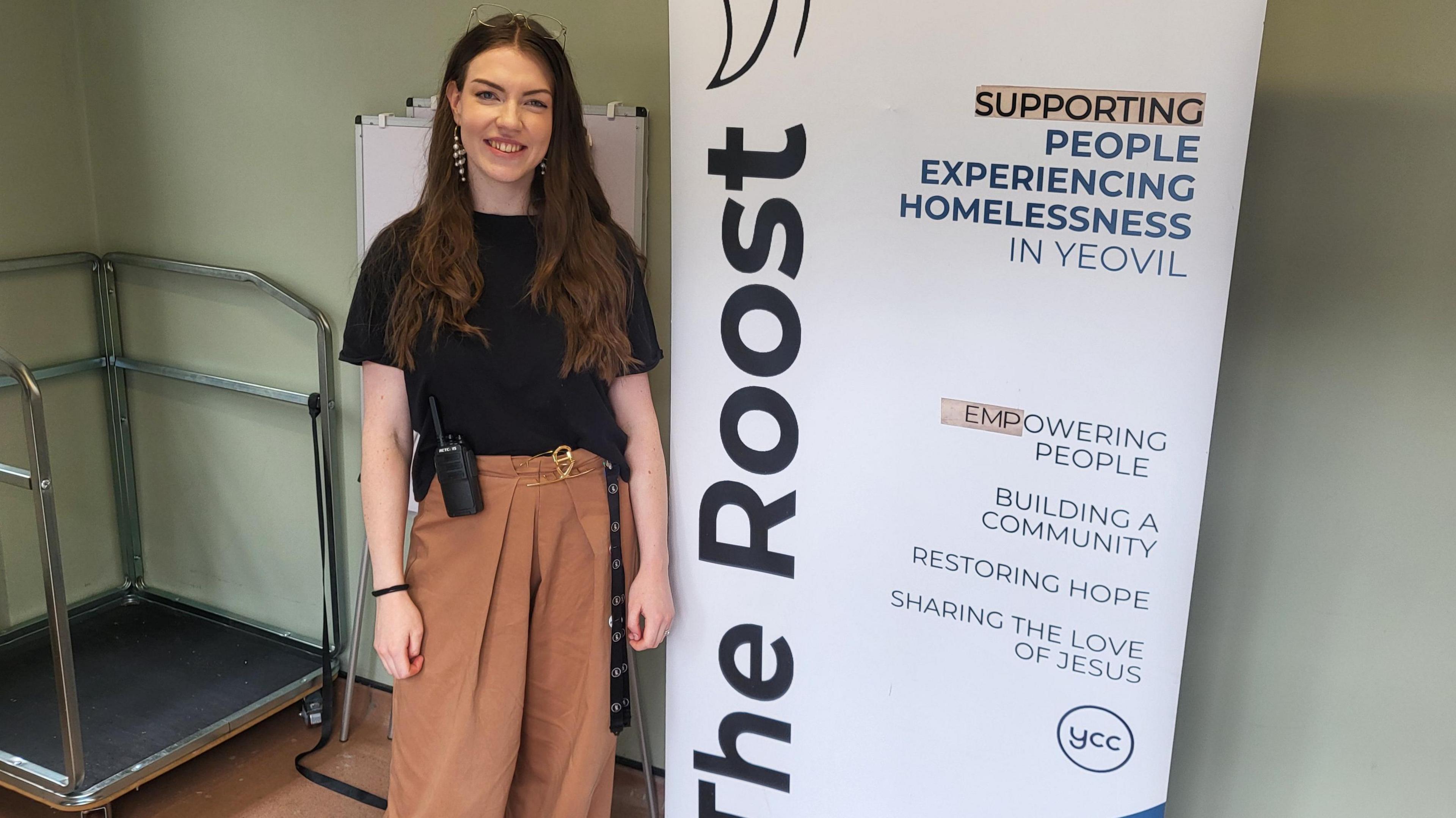Bridget Doney stood against a white sign which reads 'The Roost. Supporting people experiencing homelessness in Yeovil. Empowering people, building a community, restoring hope and sharing the love of Jesus'. Bridget is smiling into the camera.