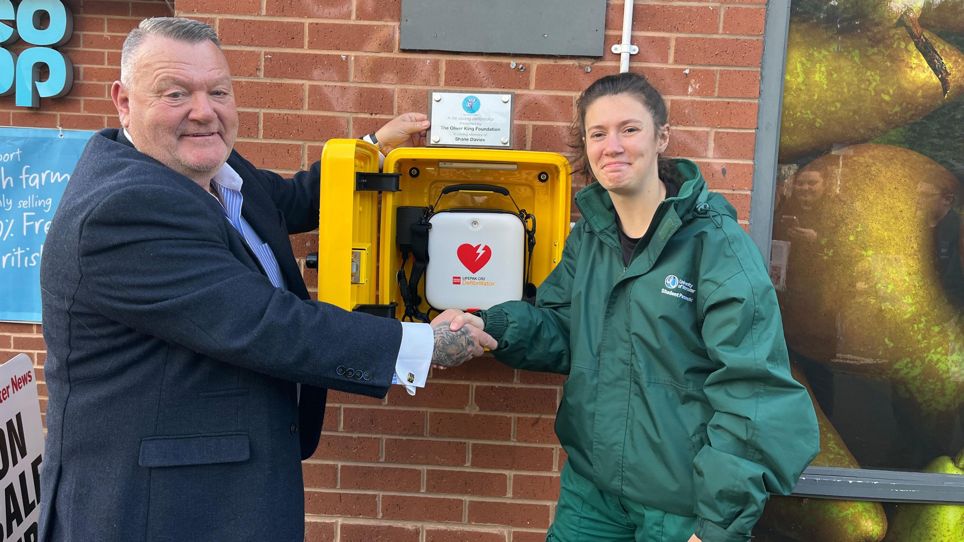 A man in a navy blue shirt with a tiger tattoo on his right hand is shaking a woman with brown hair in a ponytail and dressed in a green paramedic jacket. She is slightly smiling. Both are stood next to an opened  yellow box fixed on a red brick wall. there is a small white bag with a red heart printed on it inside.