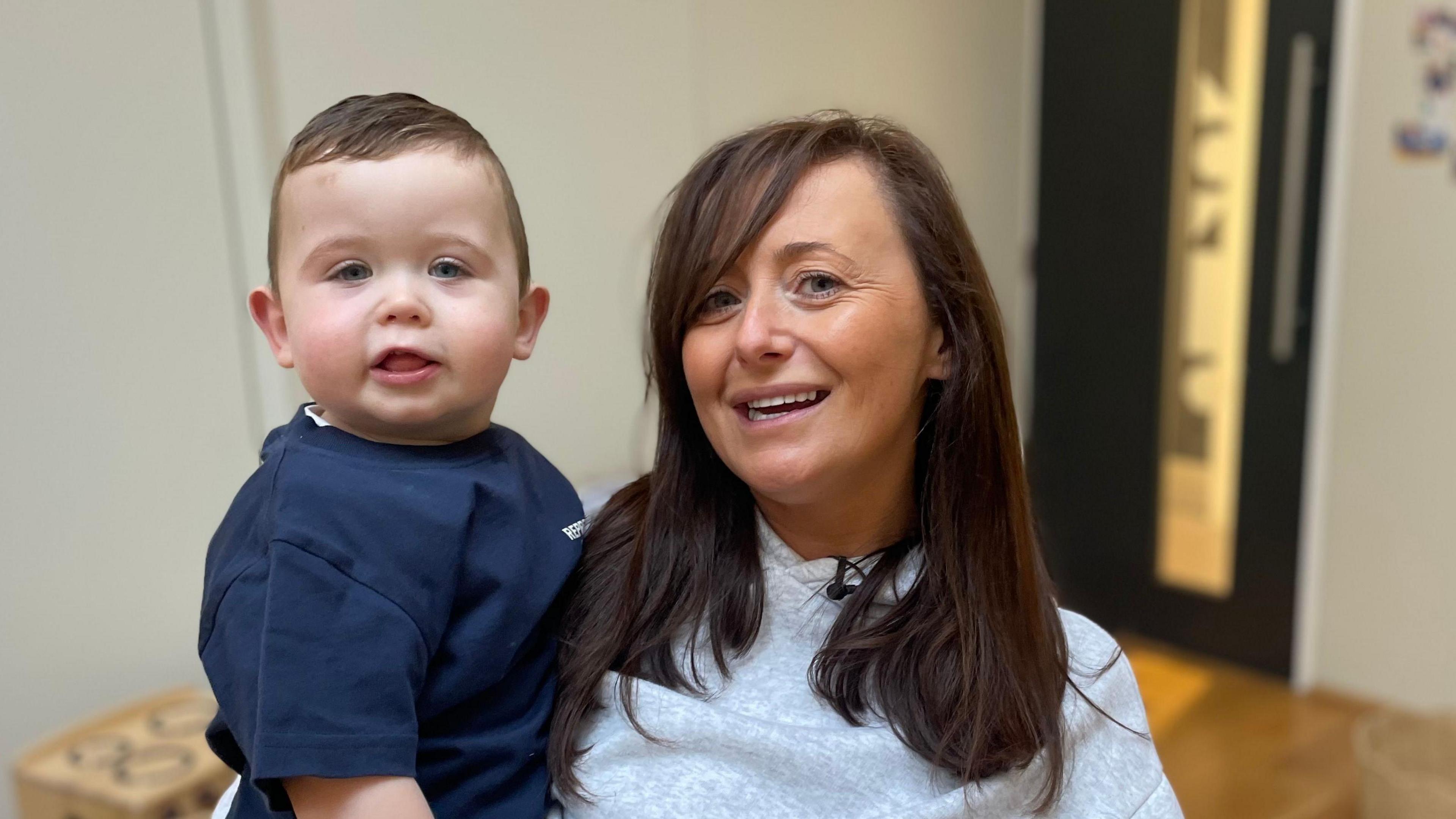 Gillian holding toddler son Frankie against her shoulder. She has lng brown hair and is wearing a grey hoodie-style jumper.  