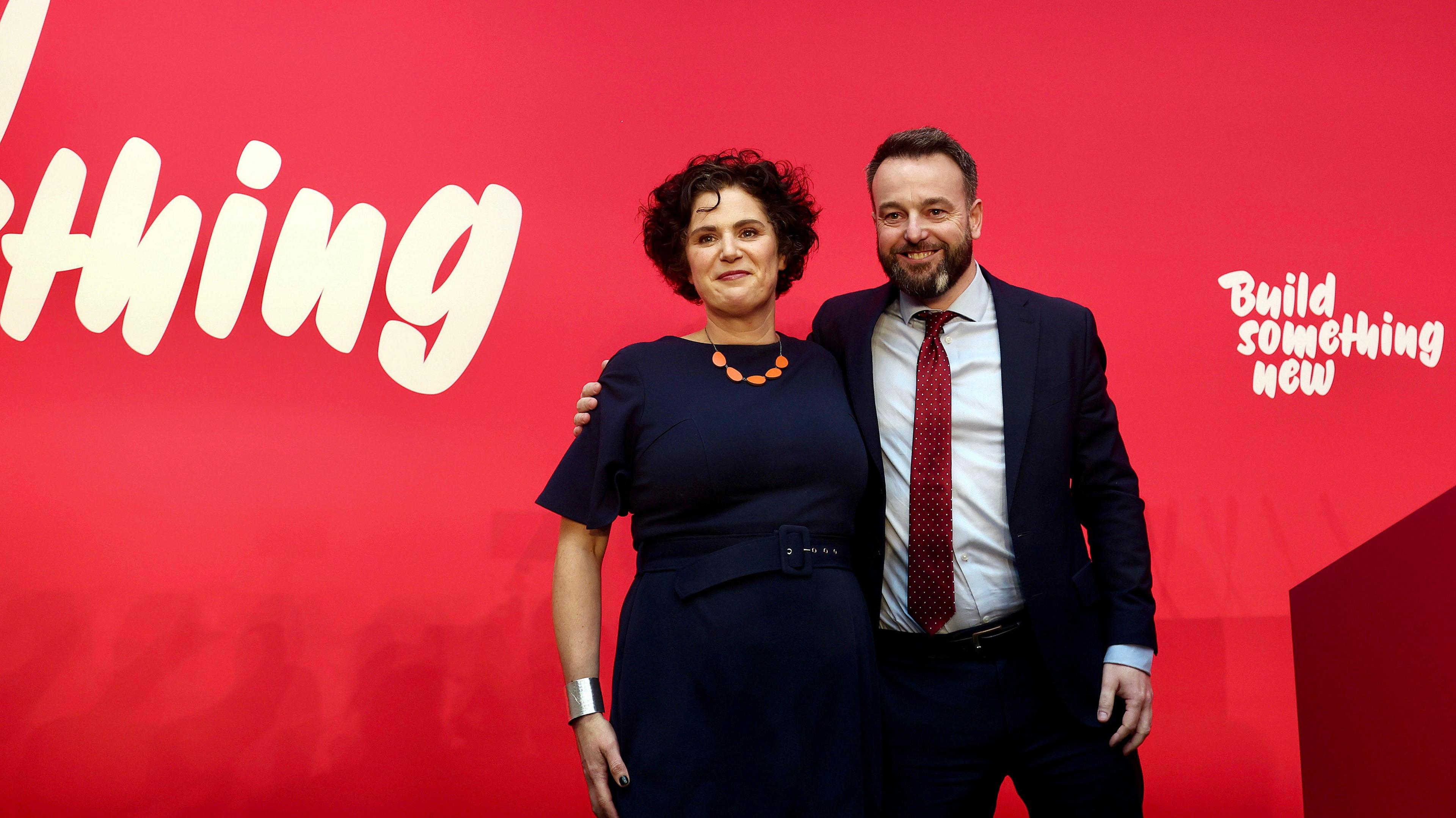 Claire Hanna and Colum Eastwood standing on stage side by side, both smiling. There is a red wall behind when with "build something new" written in white.