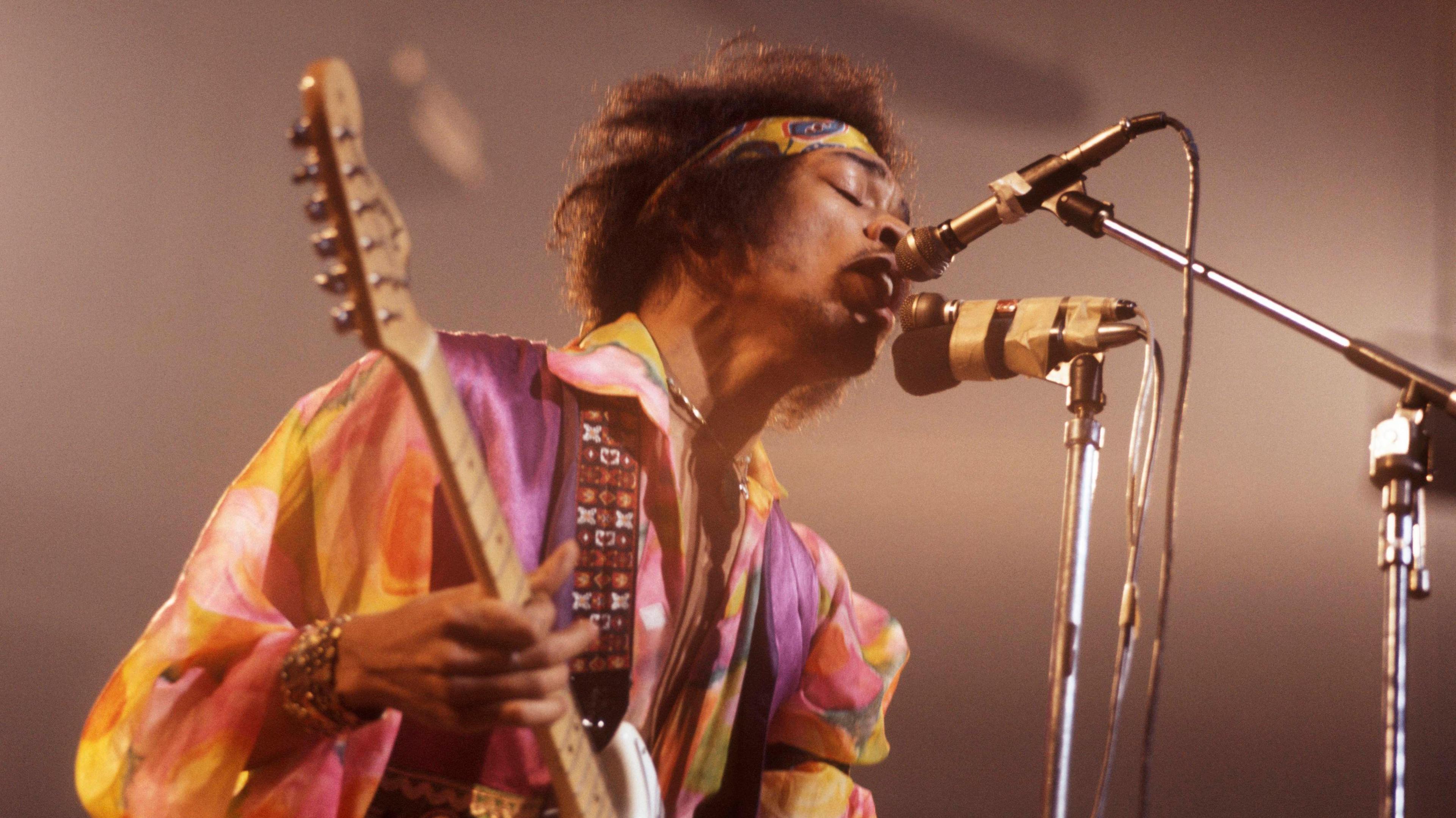 Jimi Hendrix, wearing a bright yellow shirt and purple waistcoat, with a yellow paisley bandana around his head, plays a white Fender Telecaster guitar and sings into a microphone in 1967.