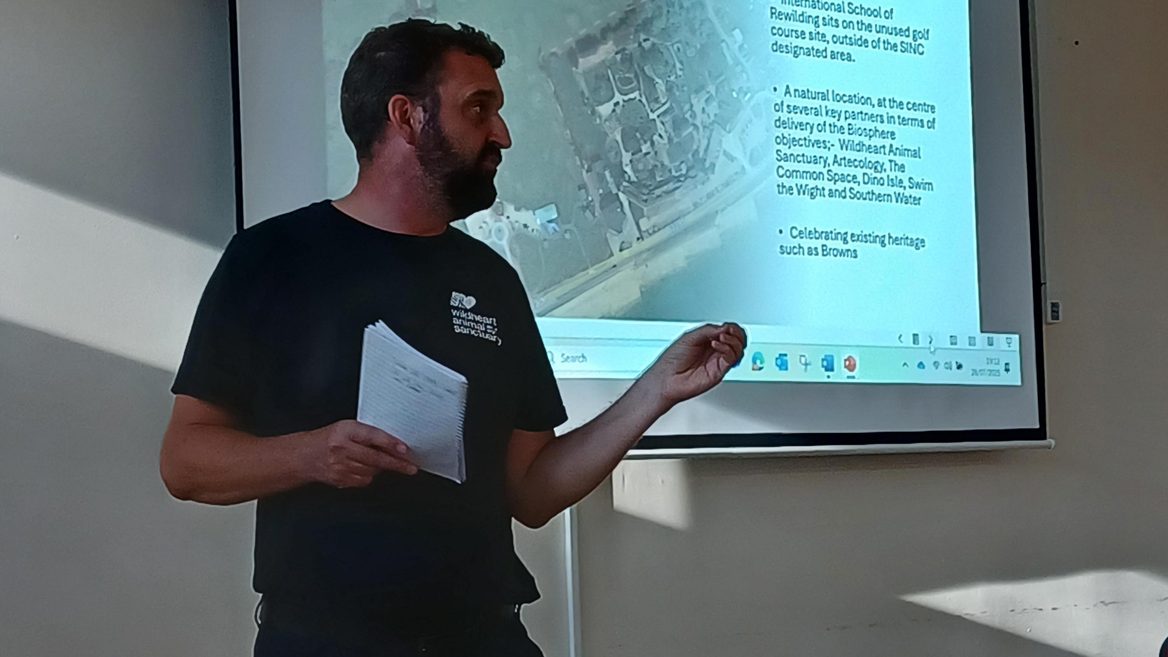 Lawrence Bates stands in a meeting room in front of a projector screen. He is looking away from the camera and holds a notepad in one hand, while his other hand is slightly reaching forward as though he is trying to emphasise his point. Mr Bates has short black hair and a short black beard. He is wearing dark trousers a short sleeved cotton t-shirt which is black and has the Wildheart Animal Sanctuary logo on the chest. 