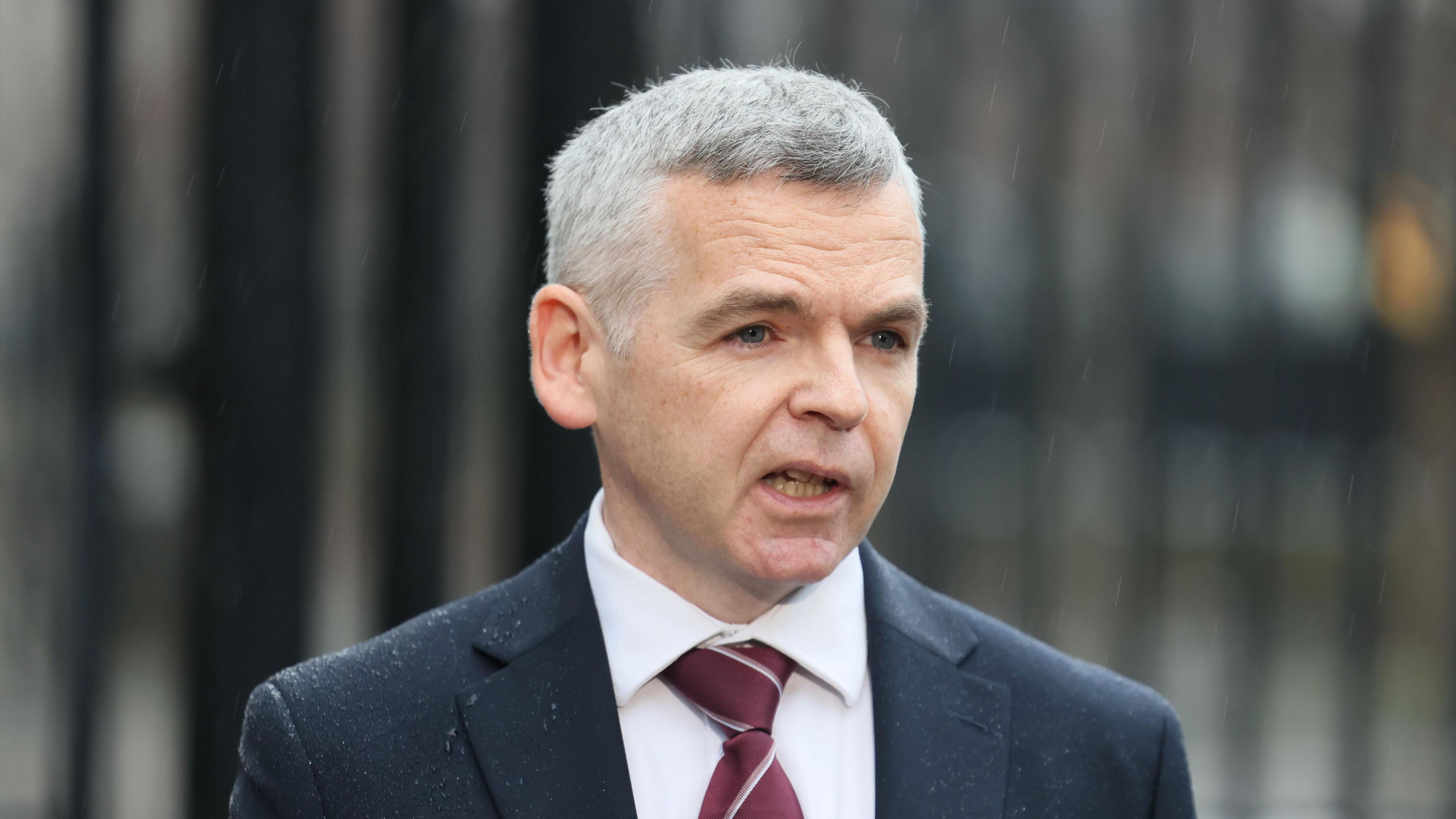 A man, with grey hair, wearing a navy suit, white shirt and burgundy tie. He is also wearing glasses and is holding a clip board and glasses. He is looking at the camera.  