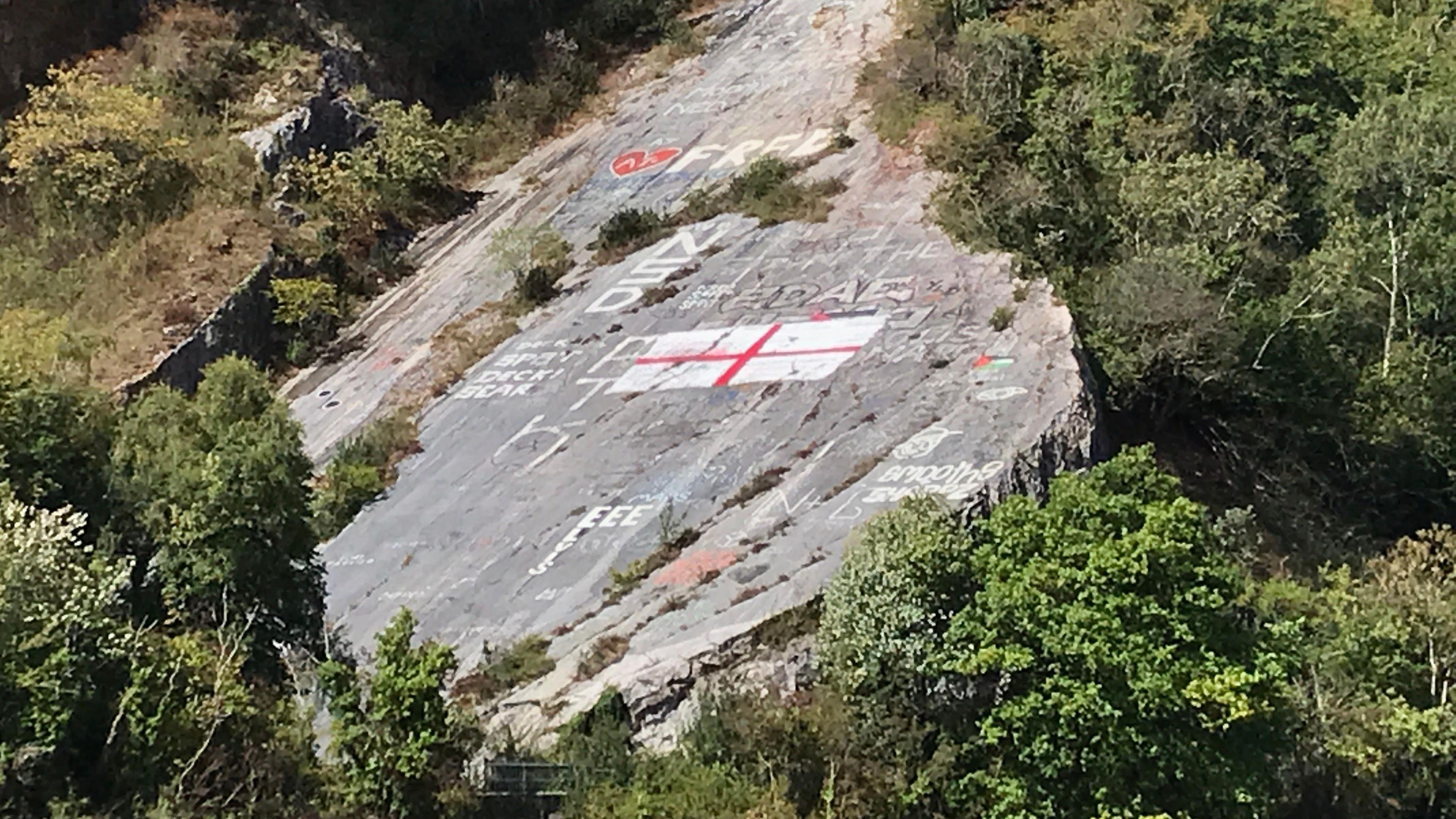 A St George's flag painted on a large sloped rock face at Avon Gorge. There is other large graffiti beside it. The rock sits amongst a lot of shrubbery.