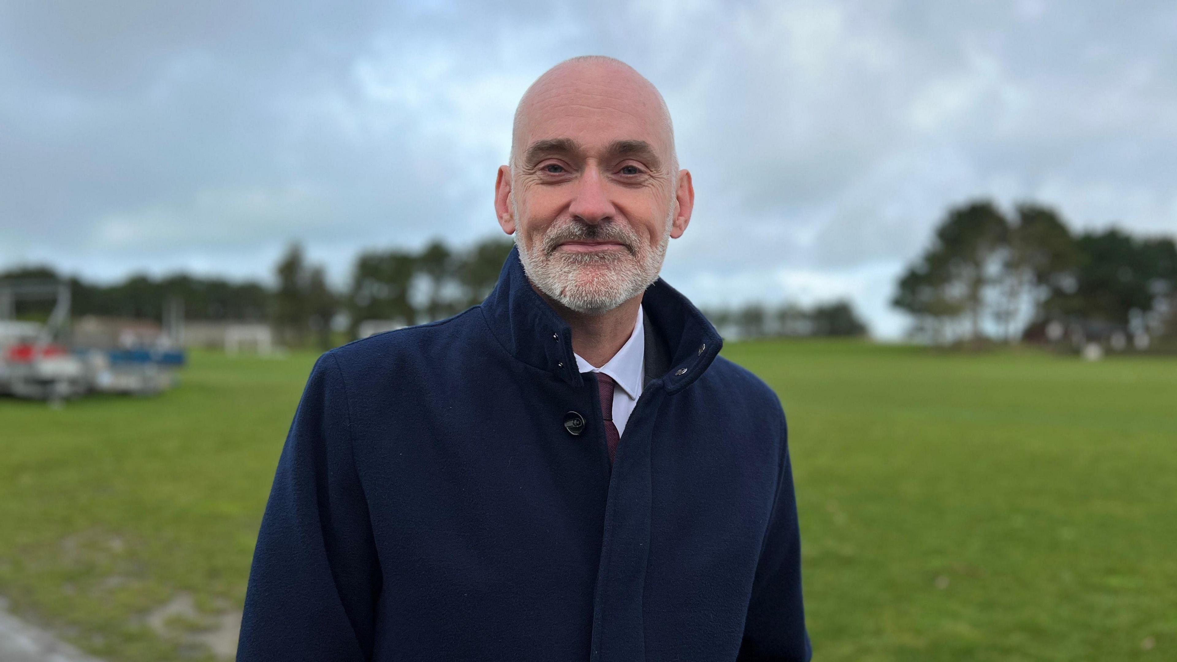 Peter Bradley - A bald man wearing a navy smart coat, a white shirt and burgundy tie. Background is blurred of a large field and trees.