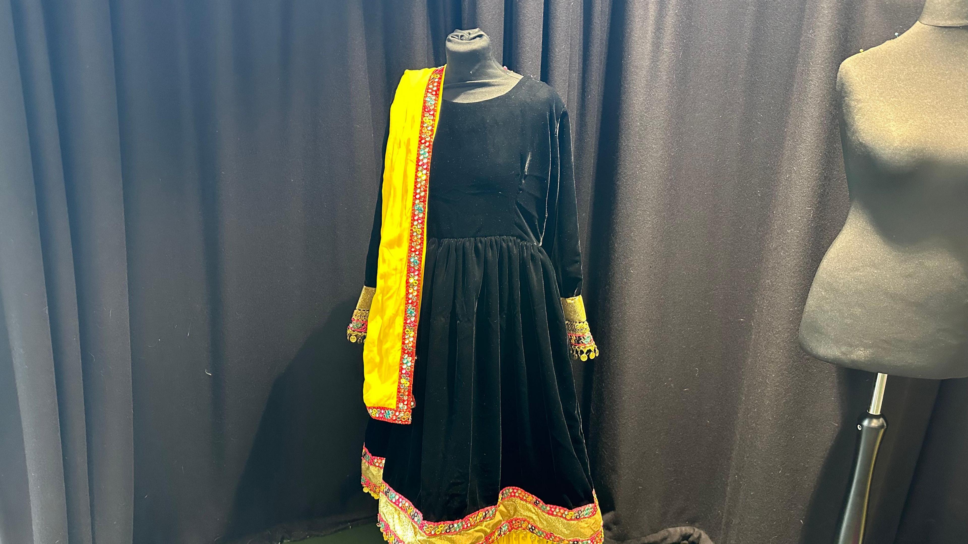 A traditional dress made by one of the group members. It is a black velvet 1950's style dress with a round neck. At the bottem there is a trim made up of a red stipe with pattens on it and a yellow plain strip which is repeated twice, there is also a matching scarf made out of the material used for the trim at the bottom