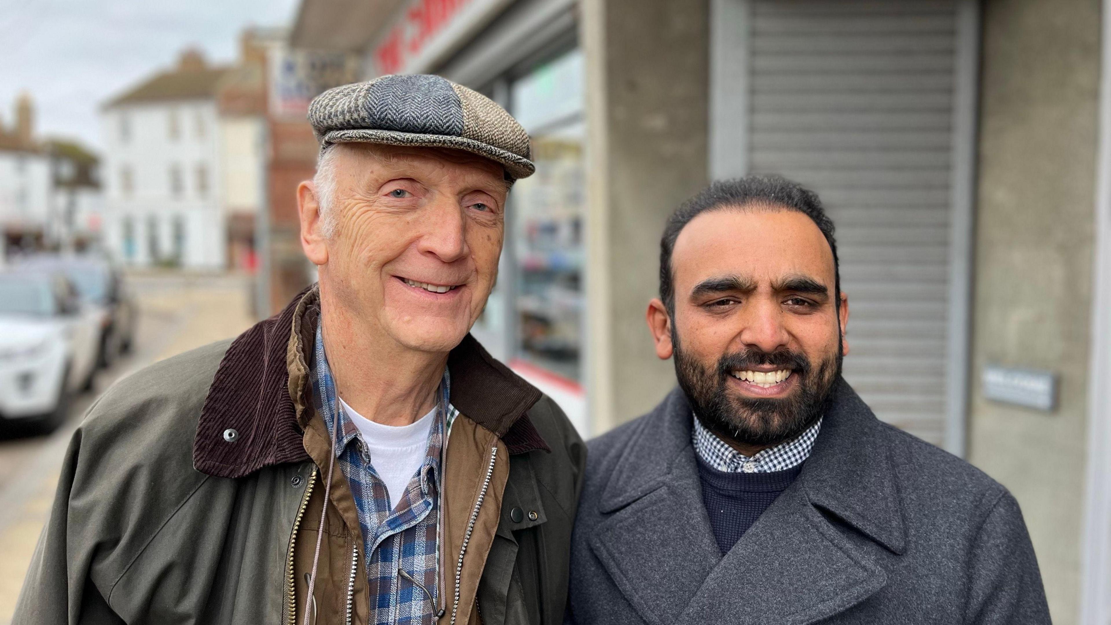 Two of the members, Anthony Joy and Khawar Shahzad, standing outside in a street smiling. Mr Joy is wearing a green jacket and flat cap, while Mr Shahzad is wearing a long wool coat, shirt and jumper.