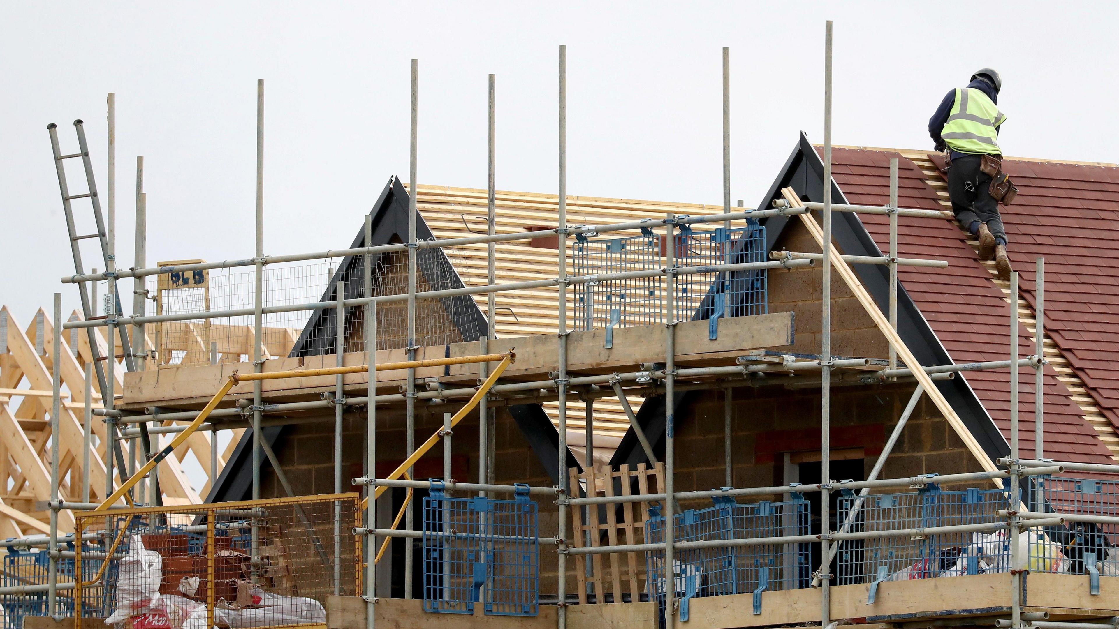 A generic shot of housebuilding, with a man tiling a roof and scaffolding against a partially built house.