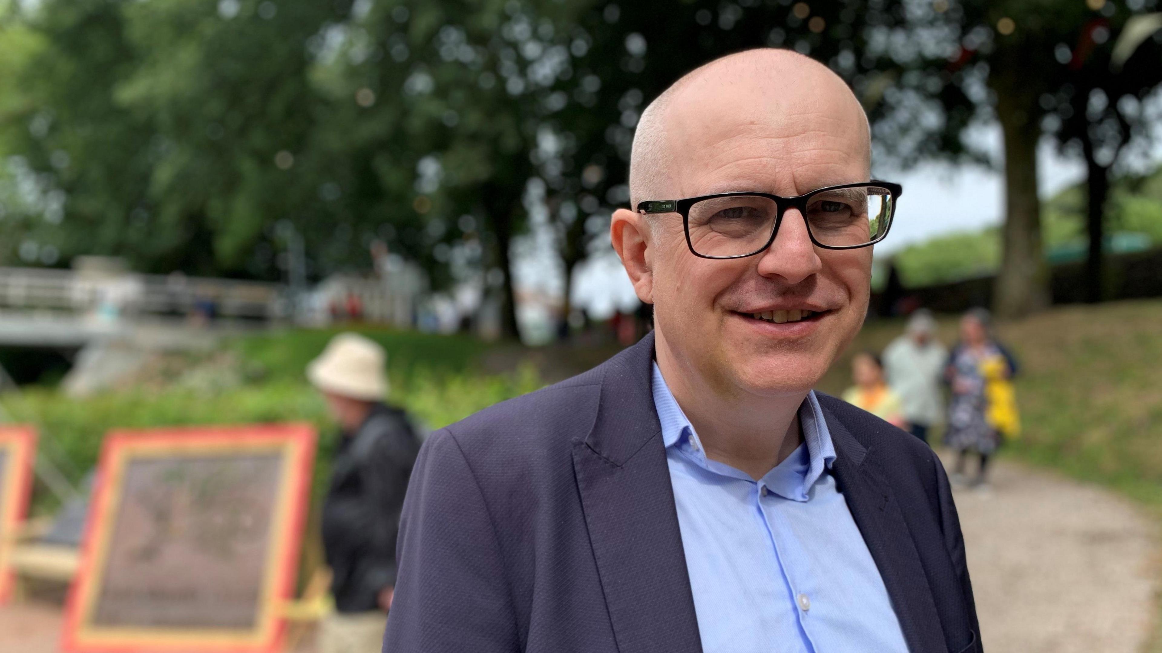 Dr Seimon Brooks, who is bald and wears glasses, wearing a blue short and suit jacket and smiling at camera, with trees and people walking in background