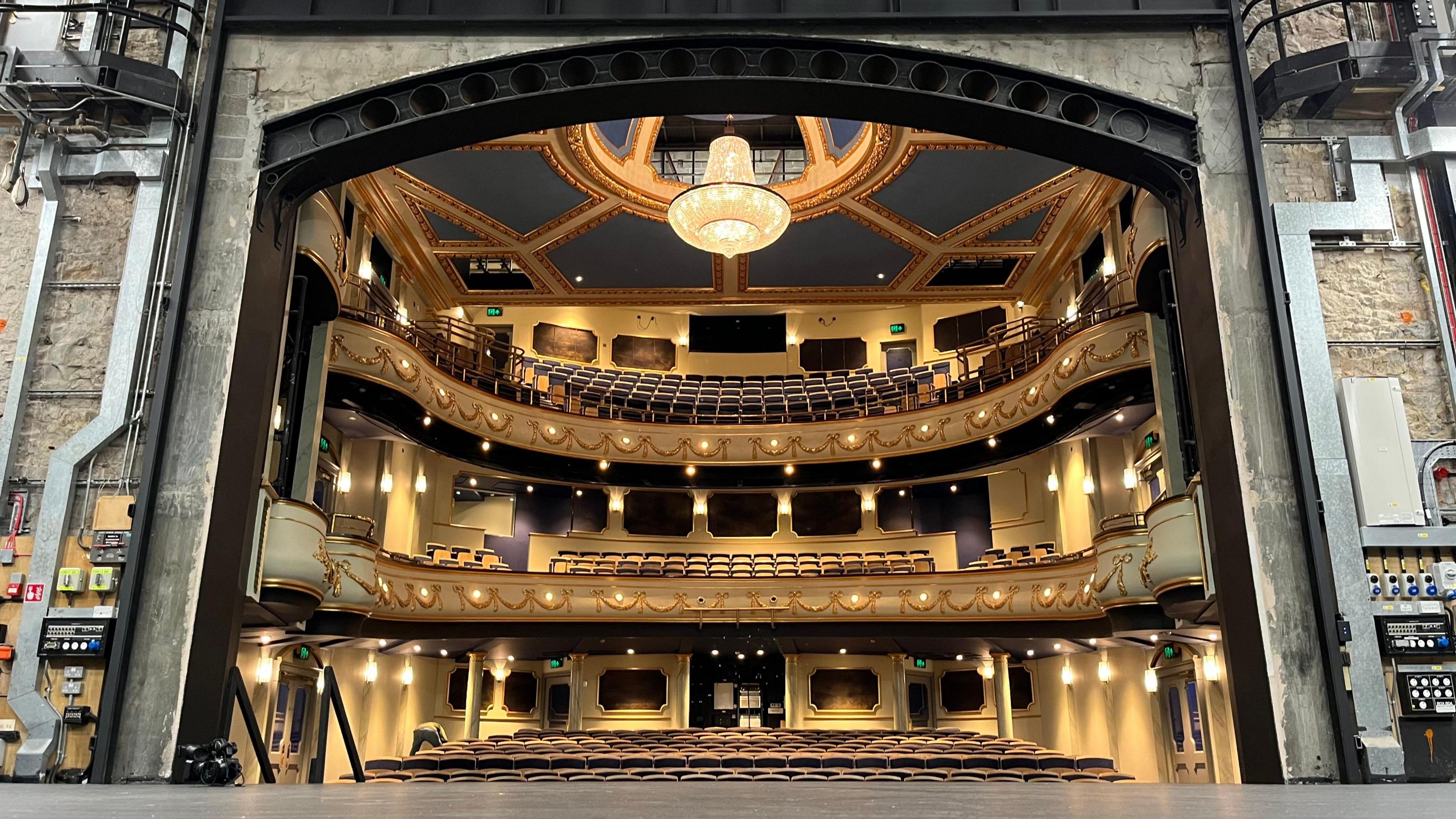 Inside Jersey Opera House. The photographer is stood on the venue's stage looking out towards the auditorium, which has gold and blue seating and walls. A large chandelier is hanging above the room.