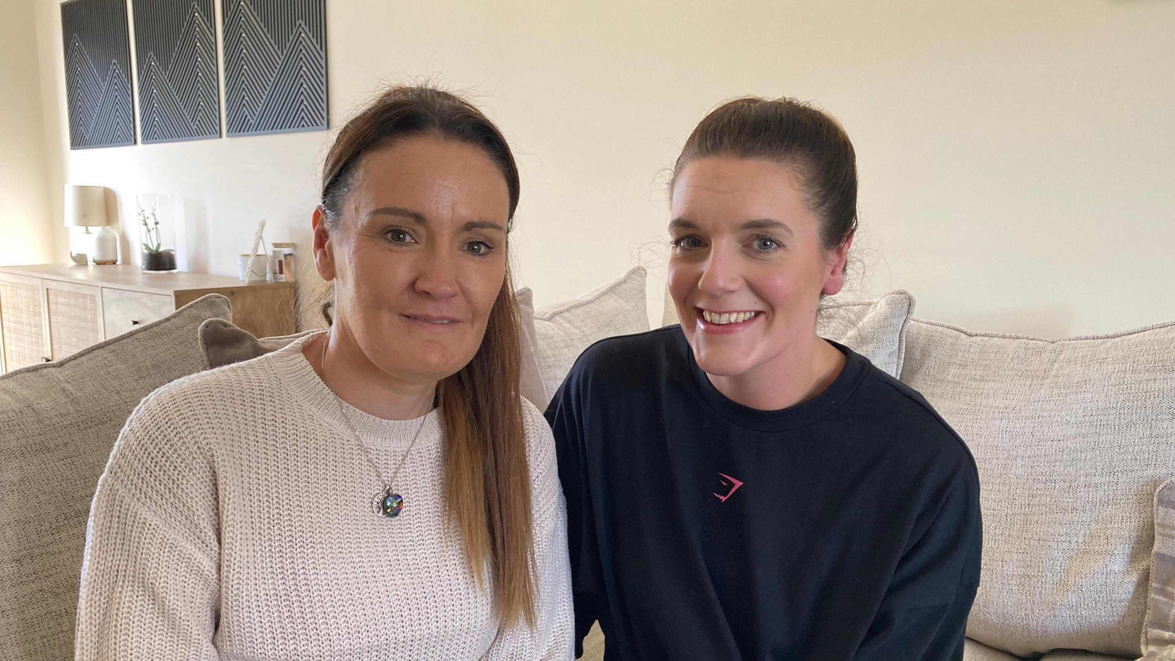 Hayley Clark, who has light brown hair and is wearing a cream sweater, and Michaela Kerr, with her brown hair up and wearing a blue sweater, sitting on a sofa inside one of their homes.