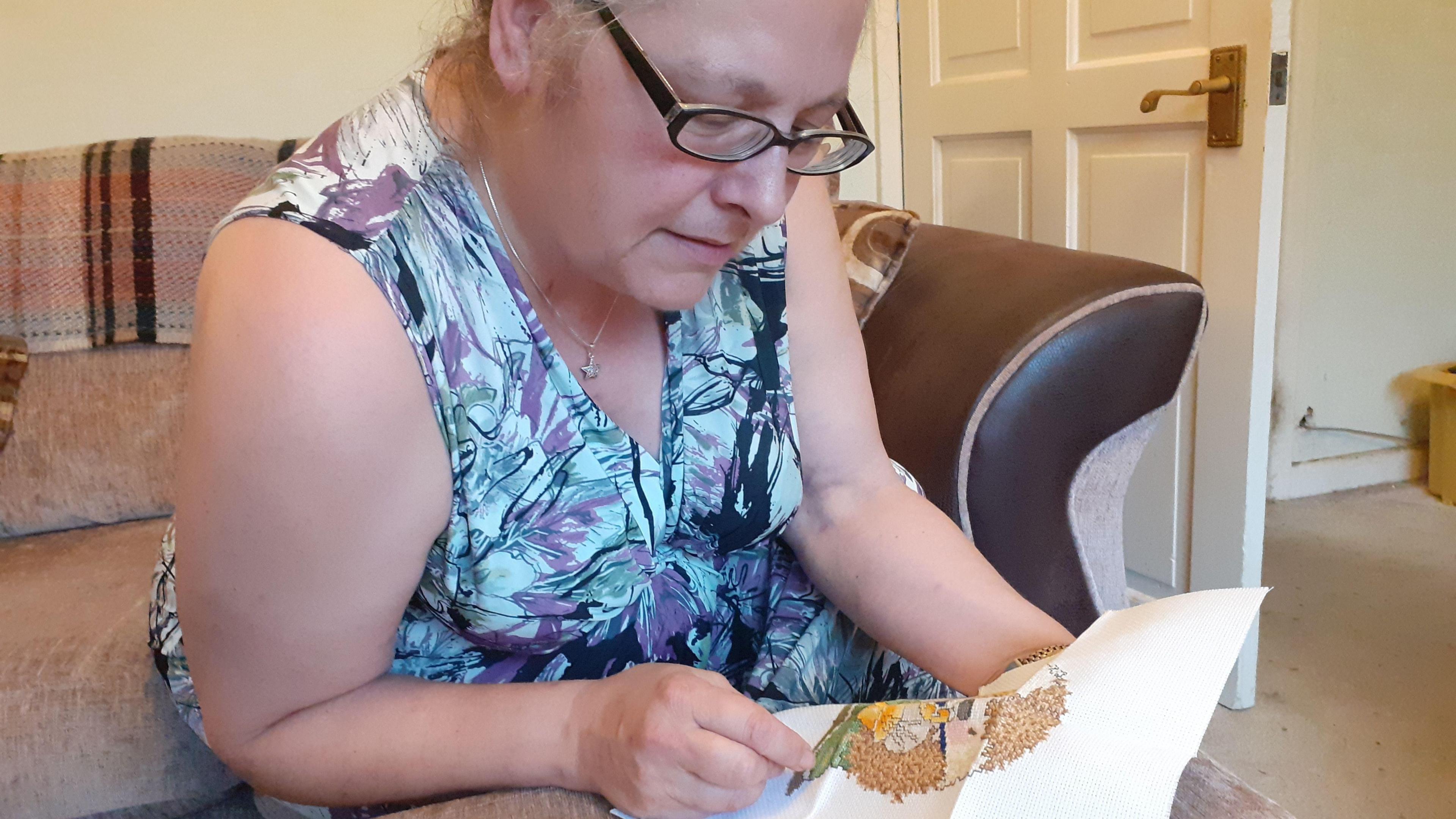 Molly Lofas, who has glasses and is wearing  a turquoise dress, working on the cross-stitch kit