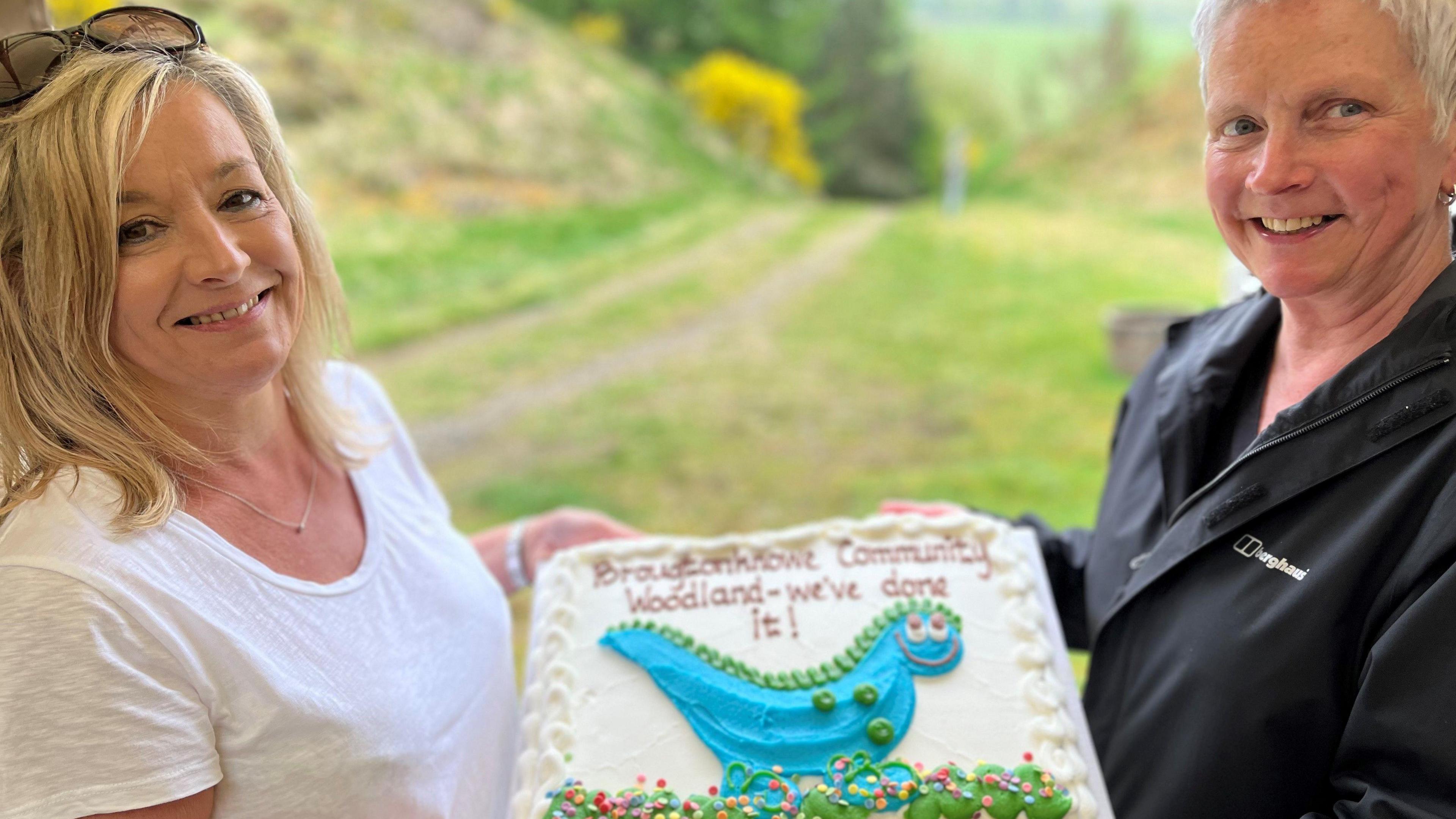 A woman with blonde hair, wearing a white T-shirt, and a grey-haired woman with a black jacket, hold a large cake with a woodland track behind them