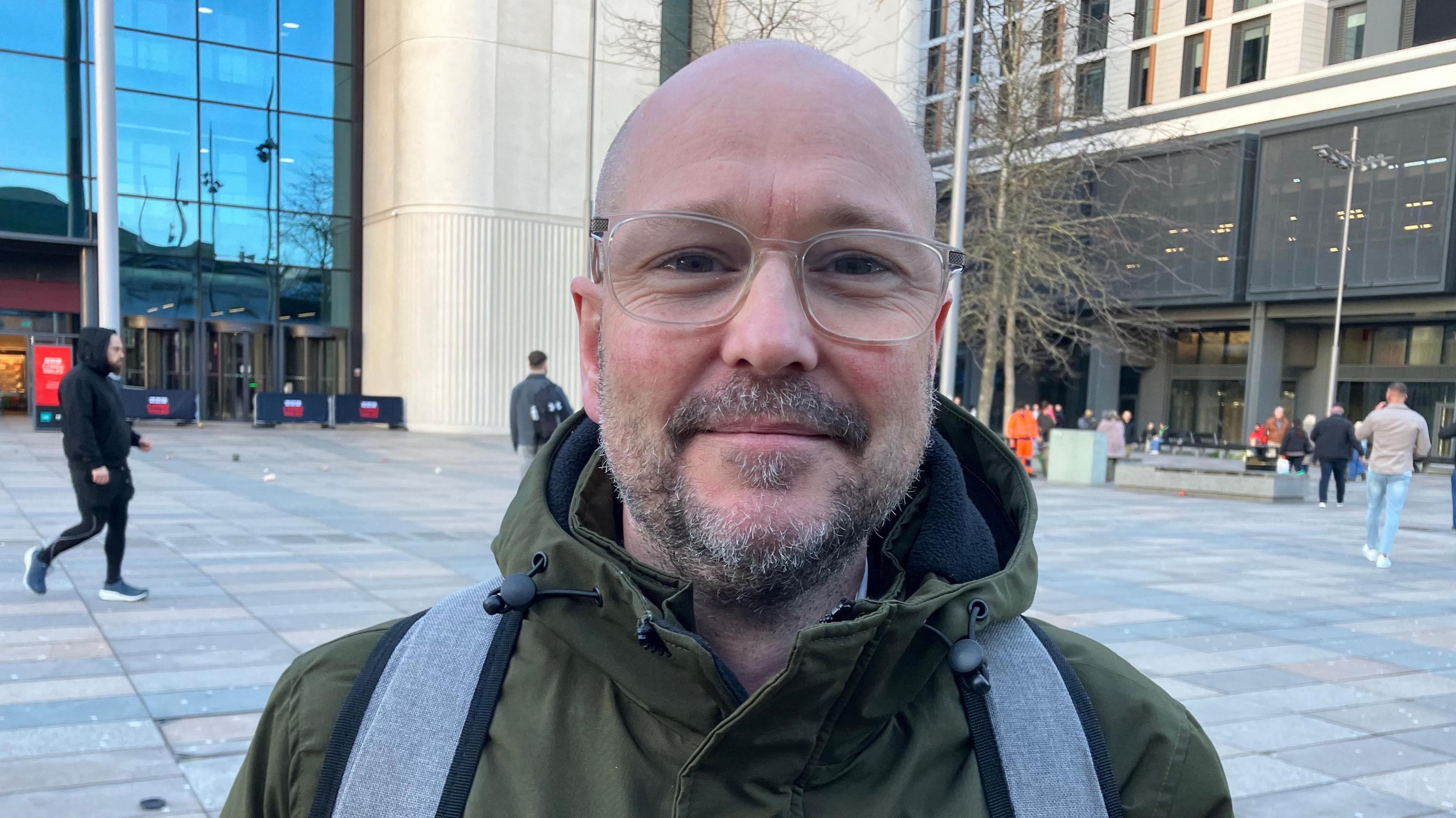 Mae Rhys Lewis yn gwisgo sbectol ac yn edrych tuag at y camera. Mae ganddo got werdd a rucksacl lwyd.