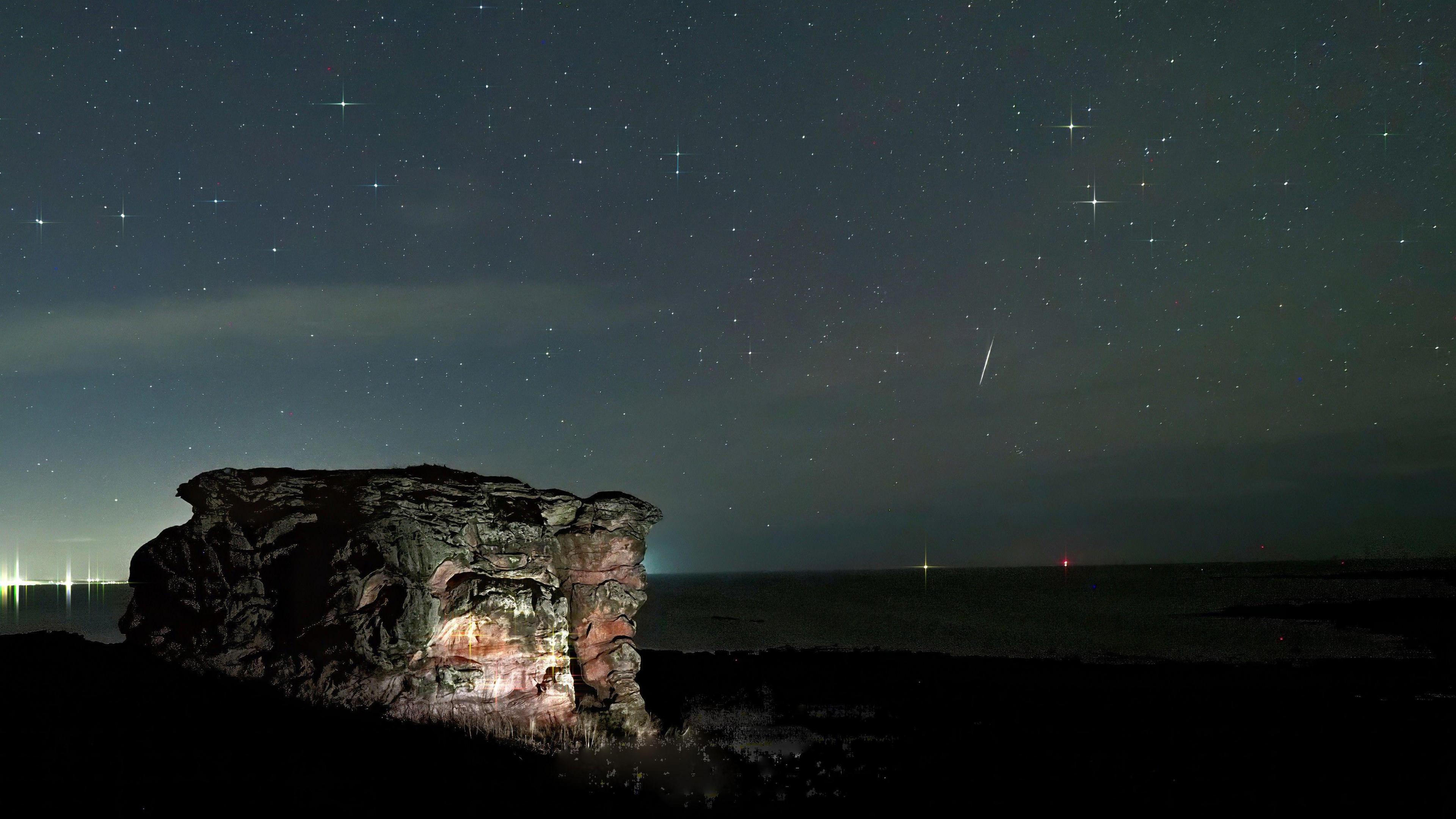 A photo showing a large rock and a starry sky at night, with a meteor showing showing some stars streaking lines across the sky