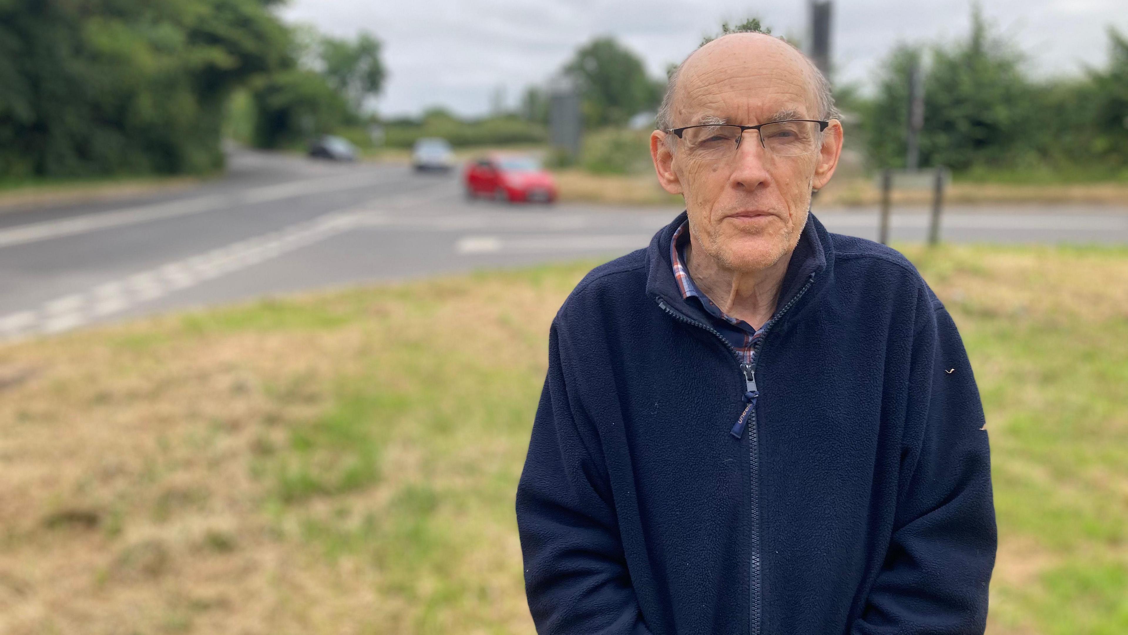 A man wearing a navy blue fleece standing on a grass verge by a road