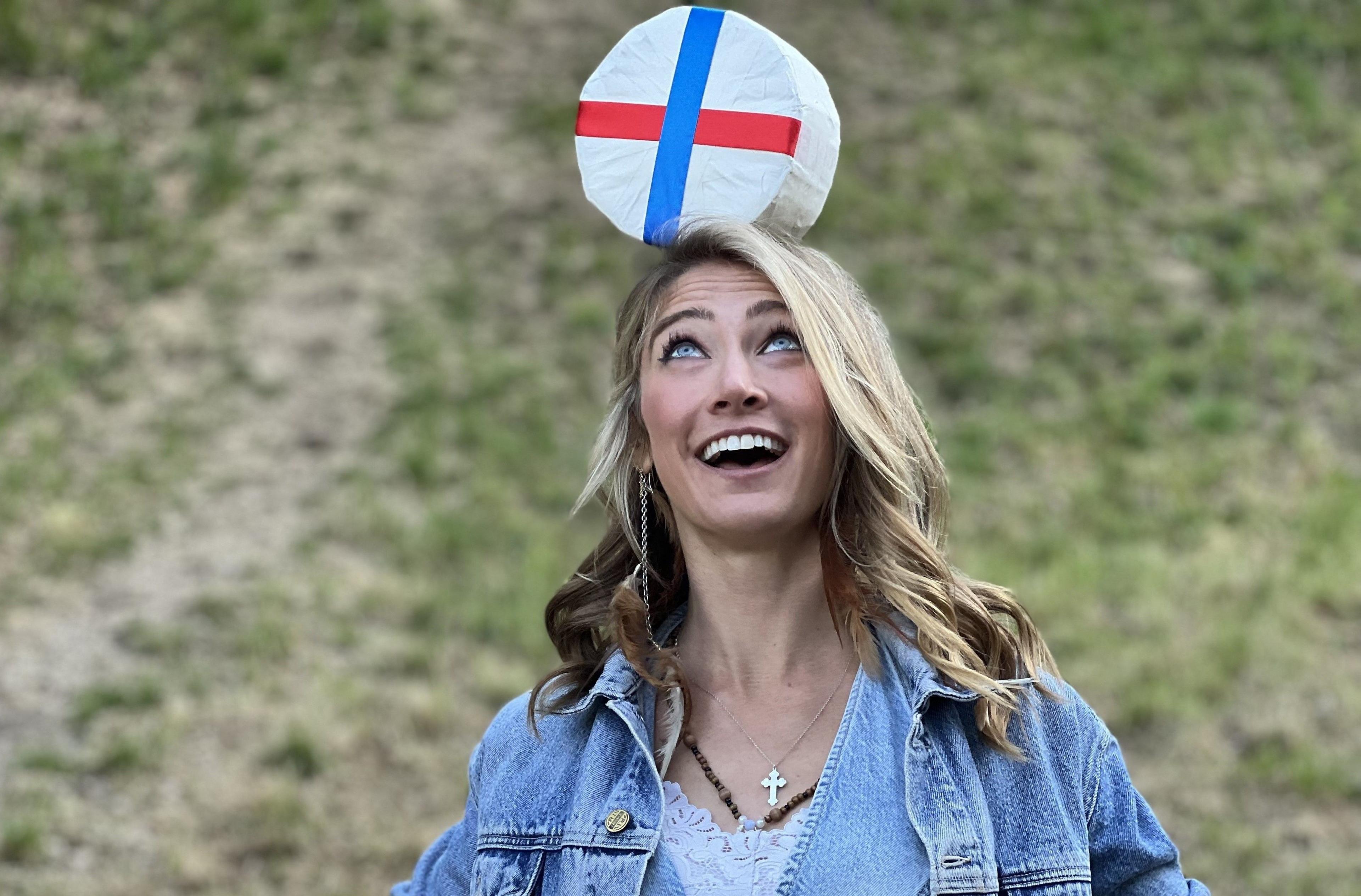 A woman with blonde hair and wearing a blue denim jacket smiles as she balances a round double gloucester cheese on her head