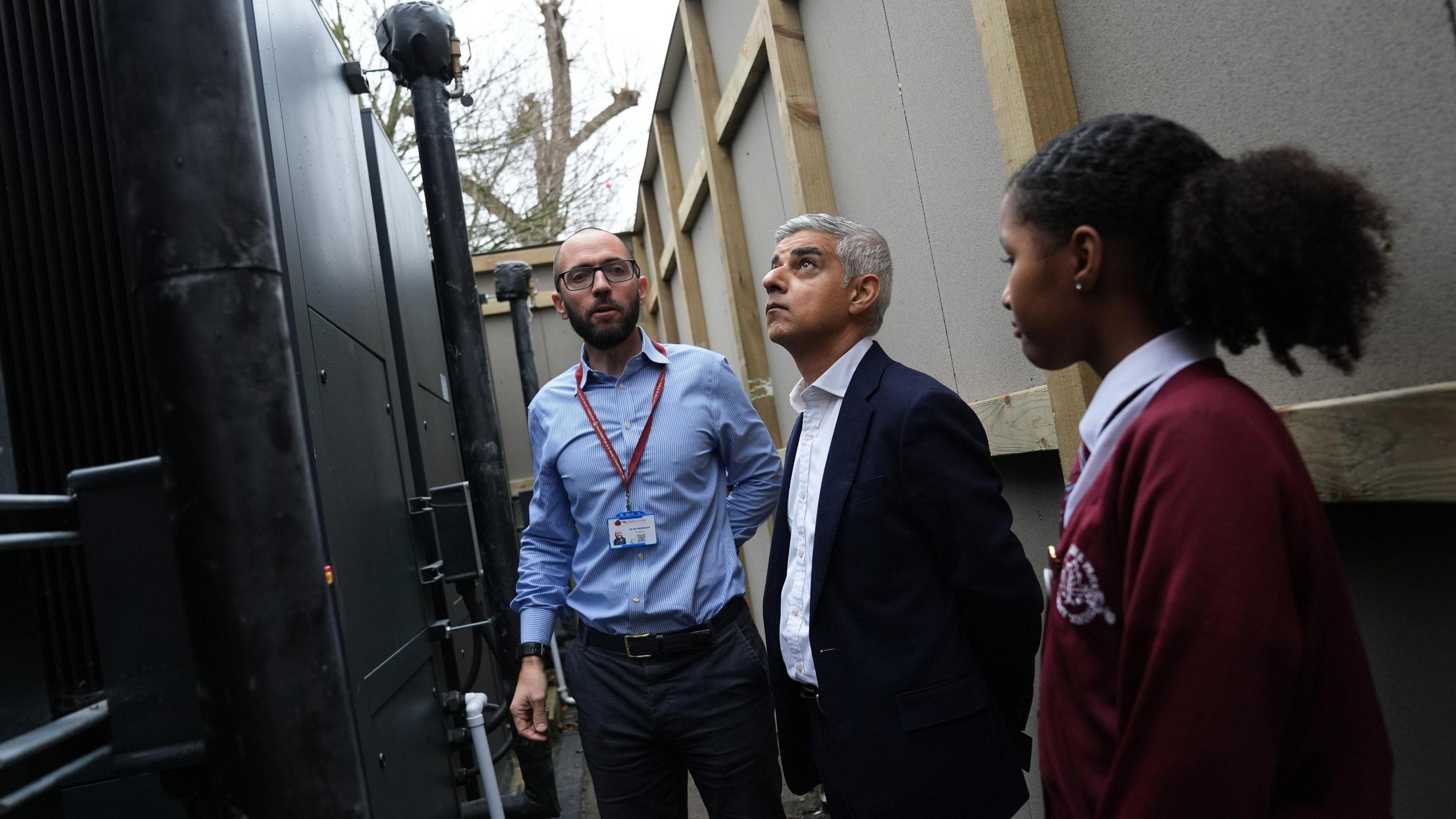 The mayor is stood looking up at a large black heat pump, alongside the headteacher who is wearing a blue shirt, a school pass and glasses and a female pupil wearing a dark red cardigan is also stood by them. 