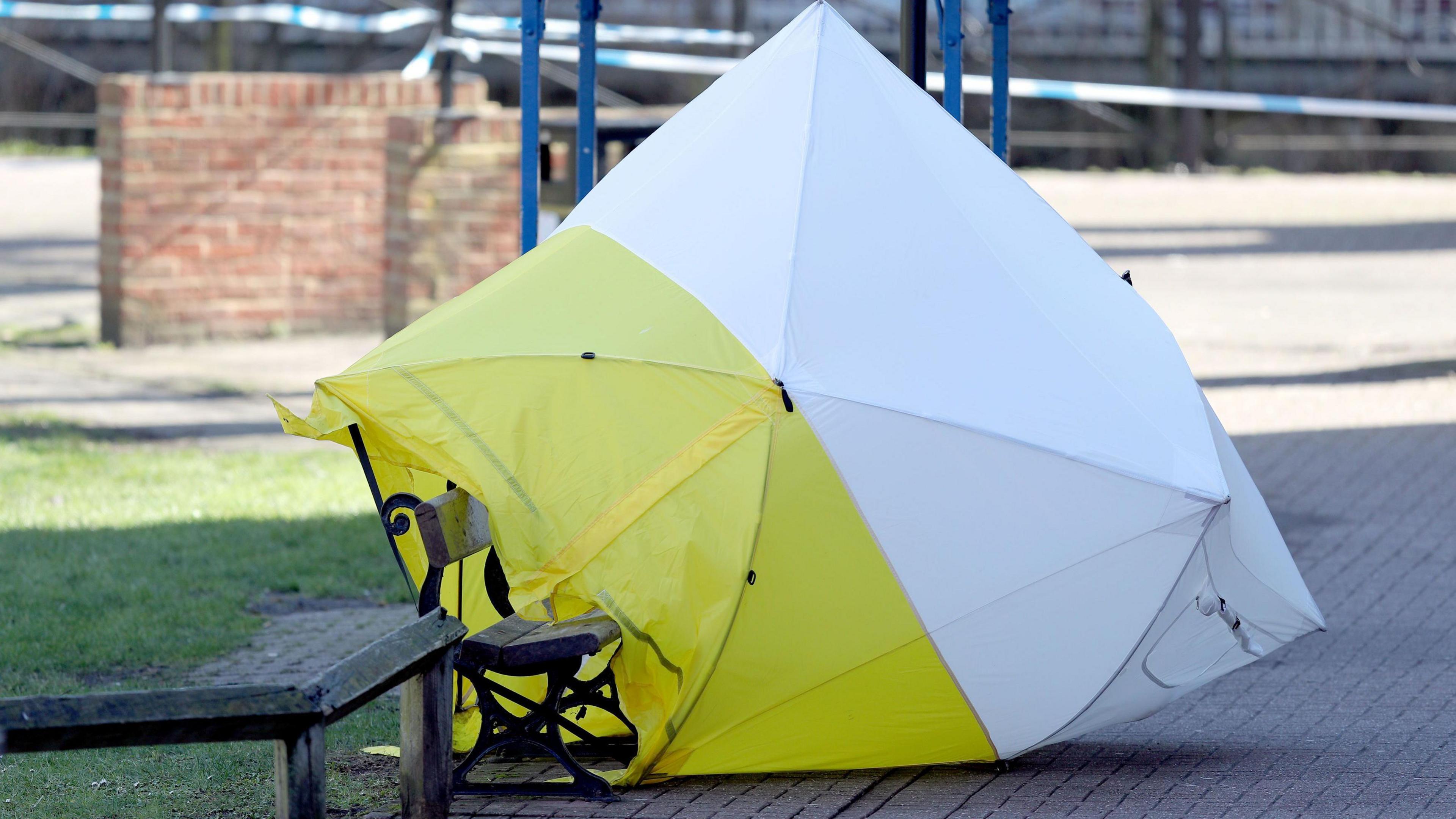 A yellow and white forensic tent covers a bench in front of a patch of grass. Police tape is visible in the background. 