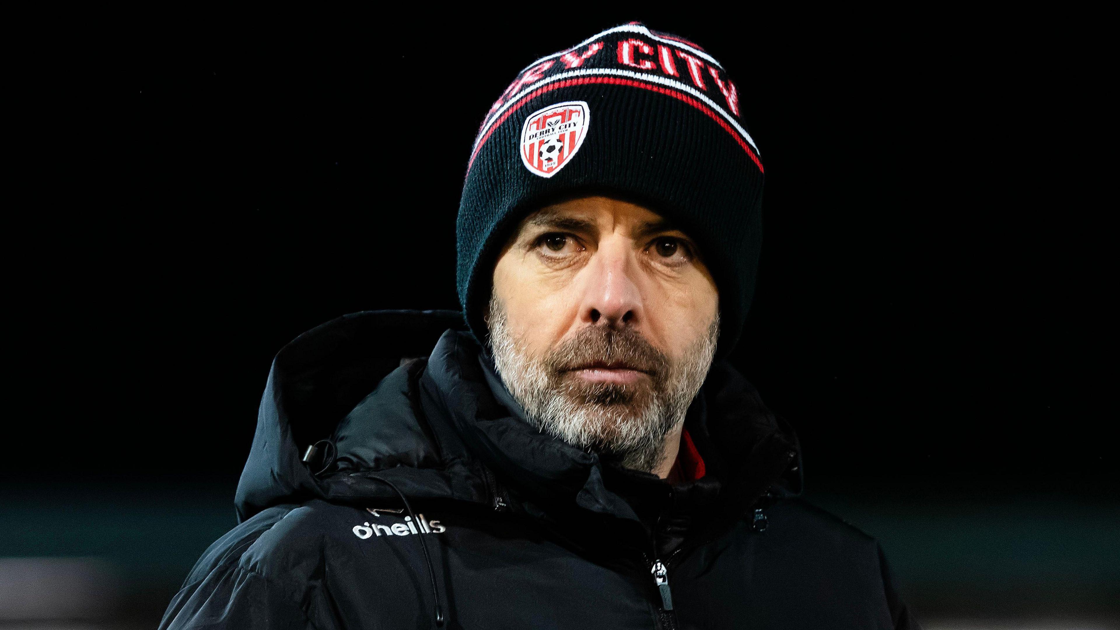 Derry City manager Tiernan Lynch speaking to the media, with a backdrop of a football stadium.