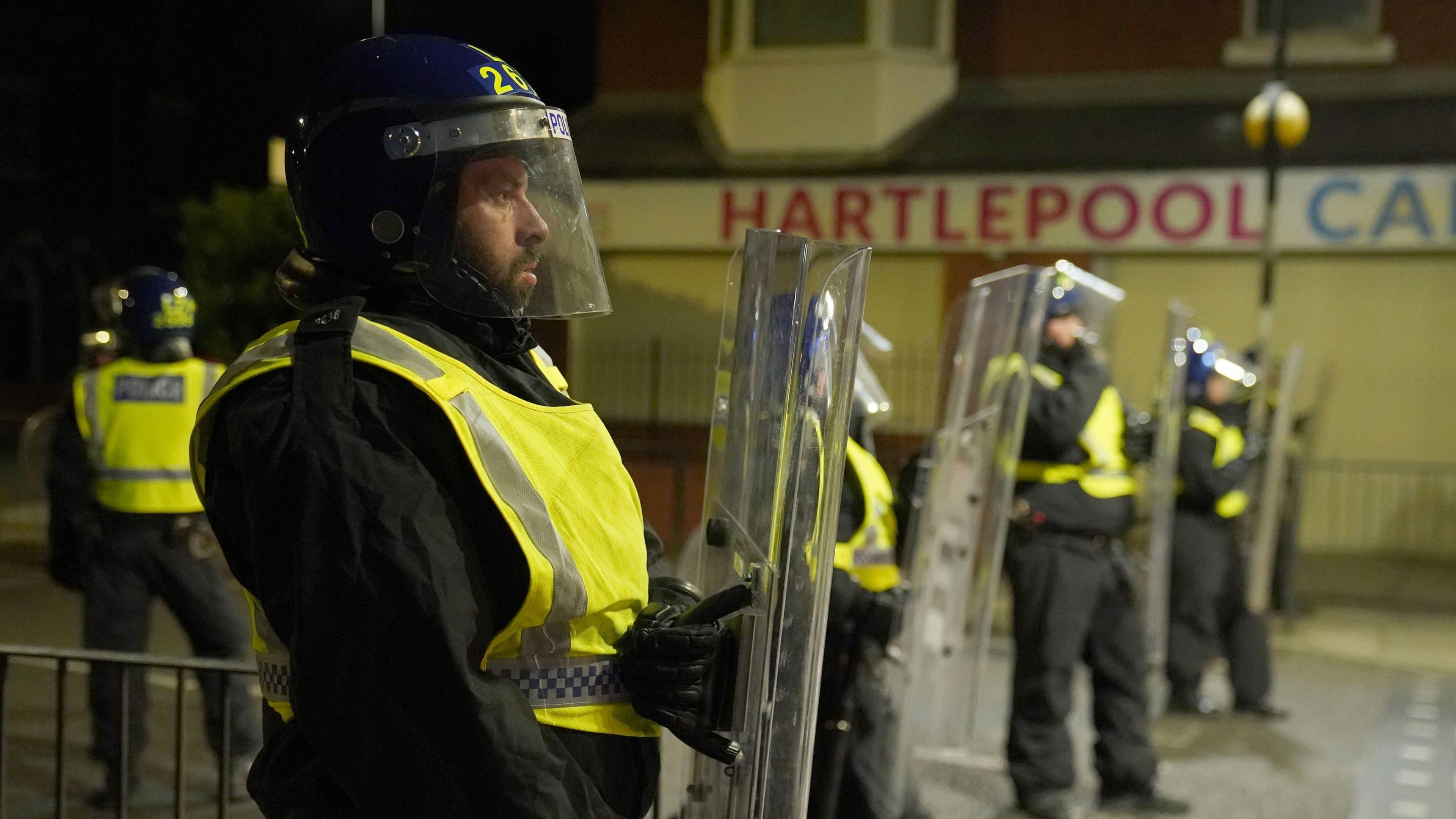 Four police officers on a street at nighttime stand in a line wearing high-vis jackets and helmets with plastic face masks, holding large plastic shields.