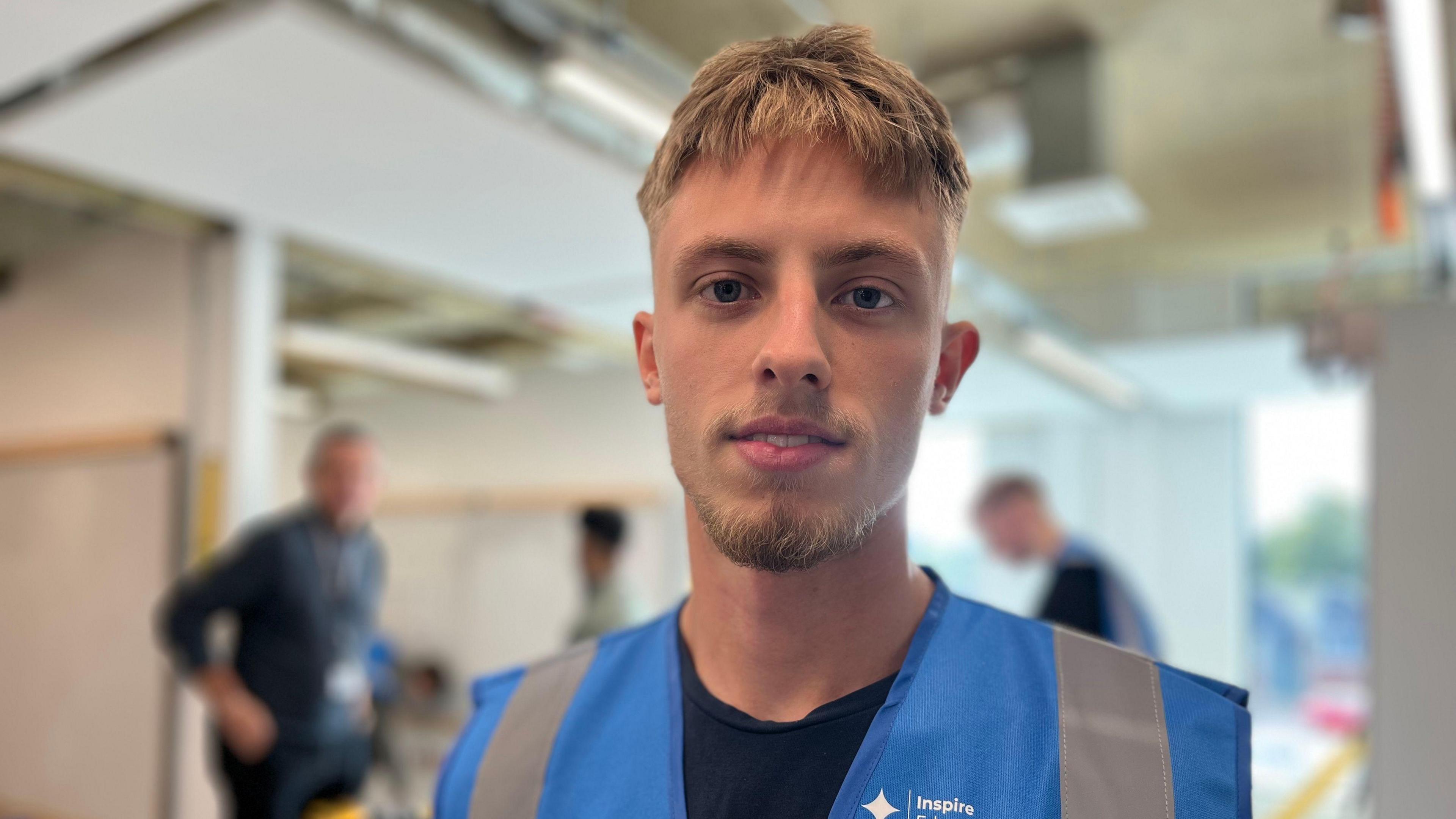 Young man with cropped hear and short beard wearing a high vis top with a workshop behind