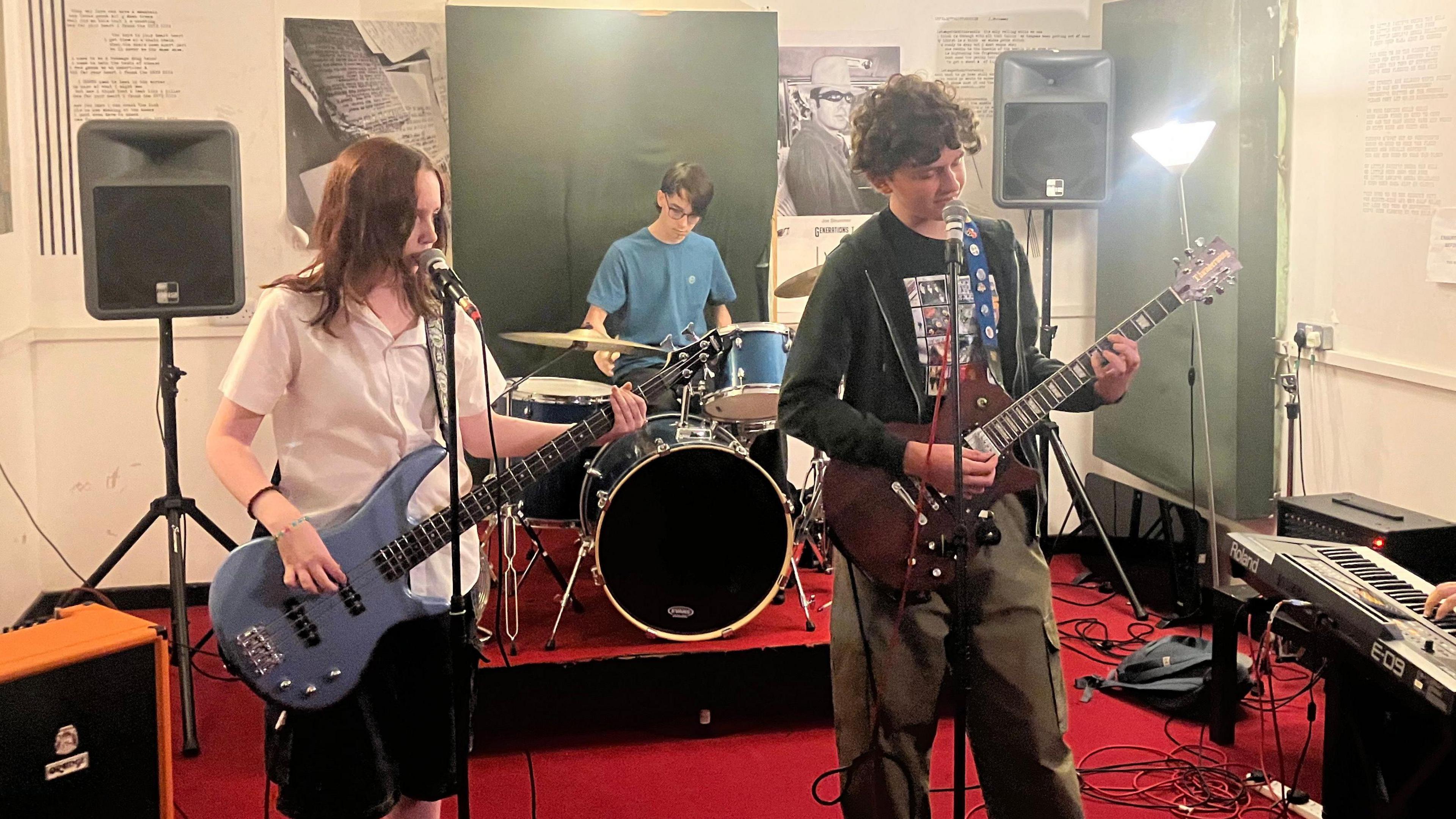 A teenage girl and boy playing guitars and singing into microphones. There is a teenage boy playing drums behind them. 