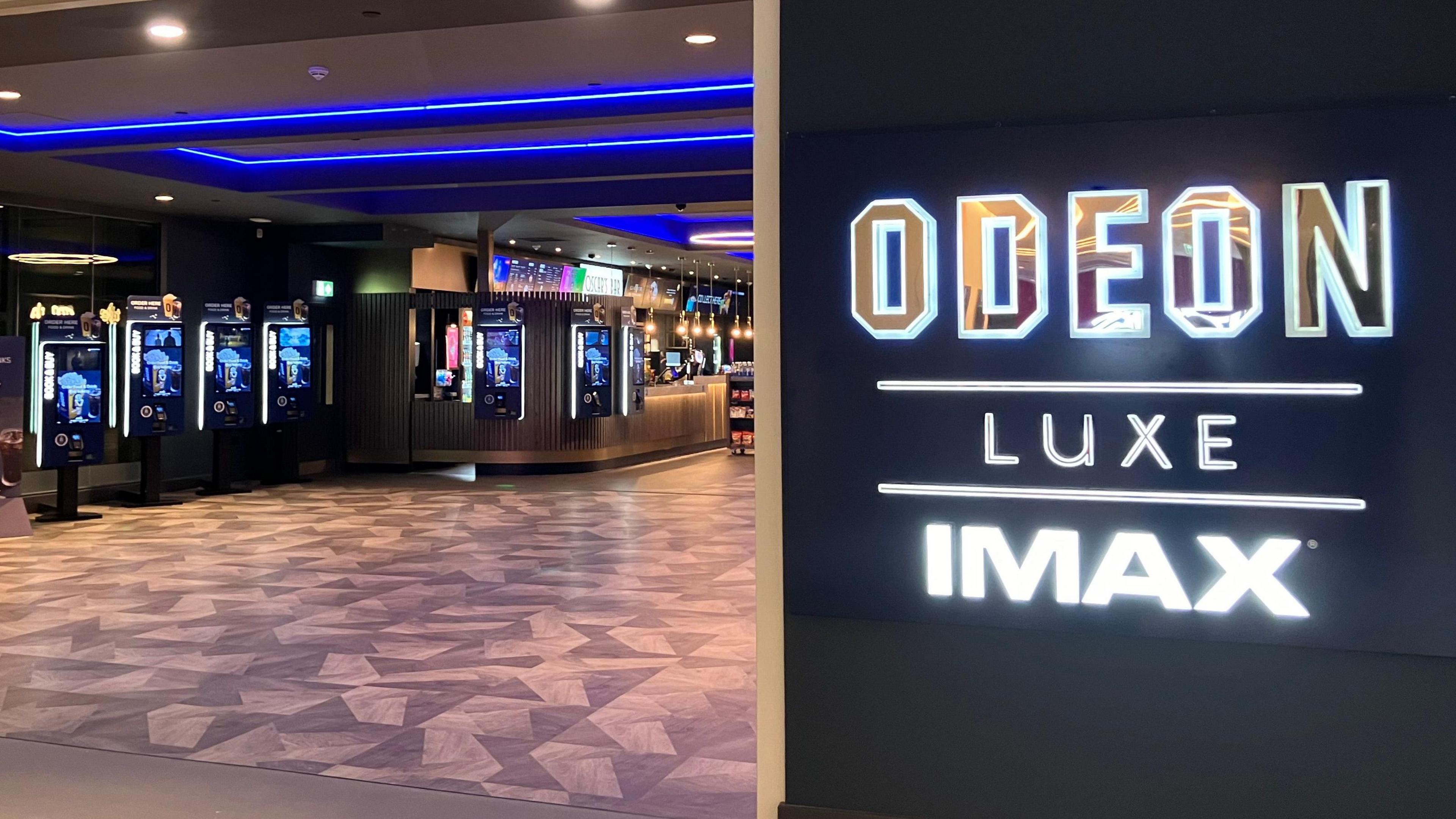 The entrance to the foyer at the new Odeon. There is a neon sign to the right that reads "Odeon Luxe IMAX". The carpet is brown with a triangular pattern. There are various drinks machines and a bar in the background. The ceiling has neon blue strips running across it.