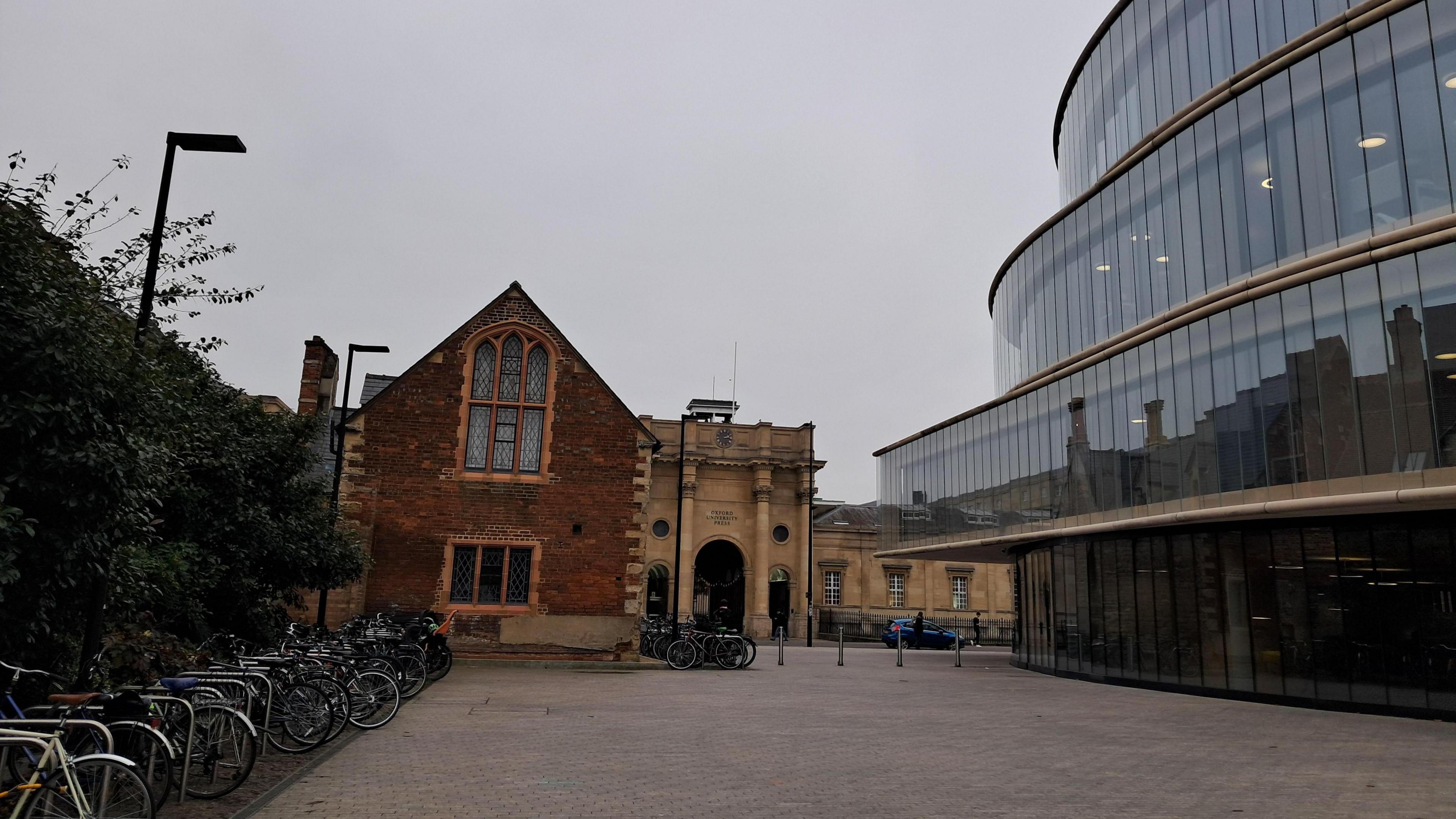 A large glass building on the right hand side of the picture reflects a brick and stone building opposite. There is a large courtyard area that has two rows of bikes in it. There is a green hedge on the left edge of the photo and the sky overhead is grey.