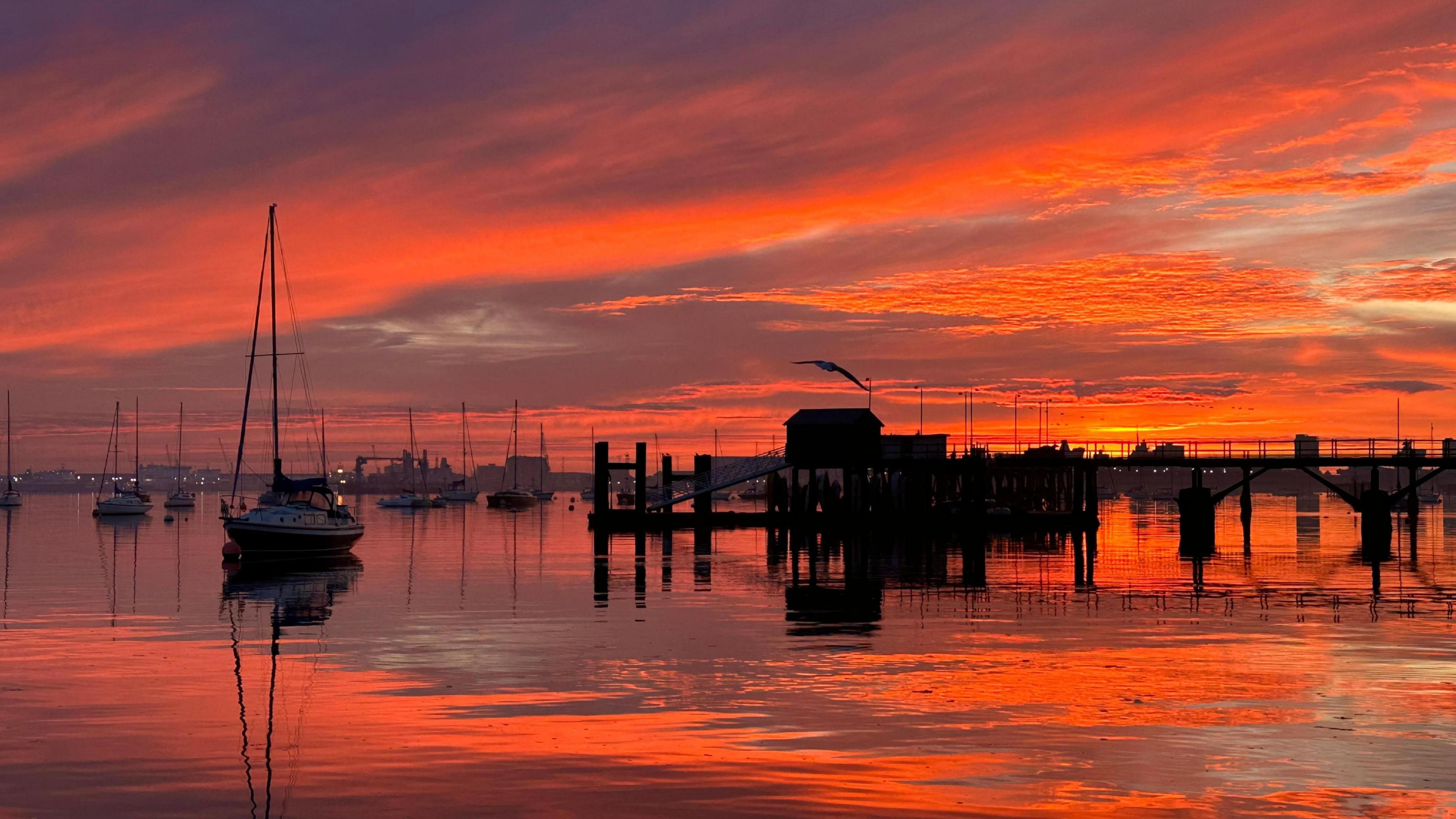 The sky glows a vivid orange over Gosport marina. There are boats moored in the water and a pier silhouetted in the background. The whole sky is glowing orange and reflecting in the water.