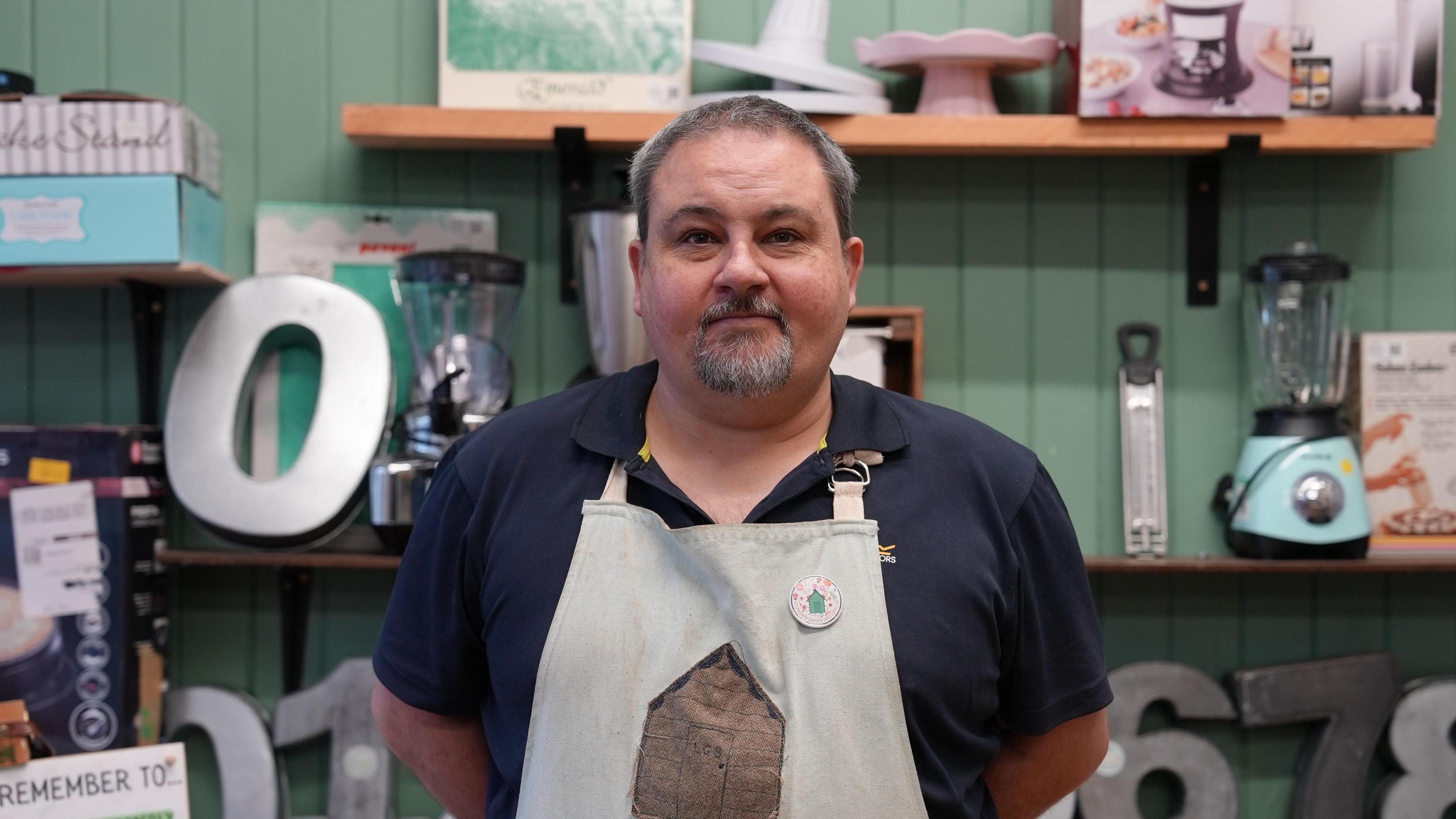 The manager of the store, Ben Mair, stands in front of the kitchen and baking section of the store. He's wearing a navy blue polo shirt and a pale blue apron with a brown shed on it. Ben has a greyish goatee and short hair. He's looking directly at the camera