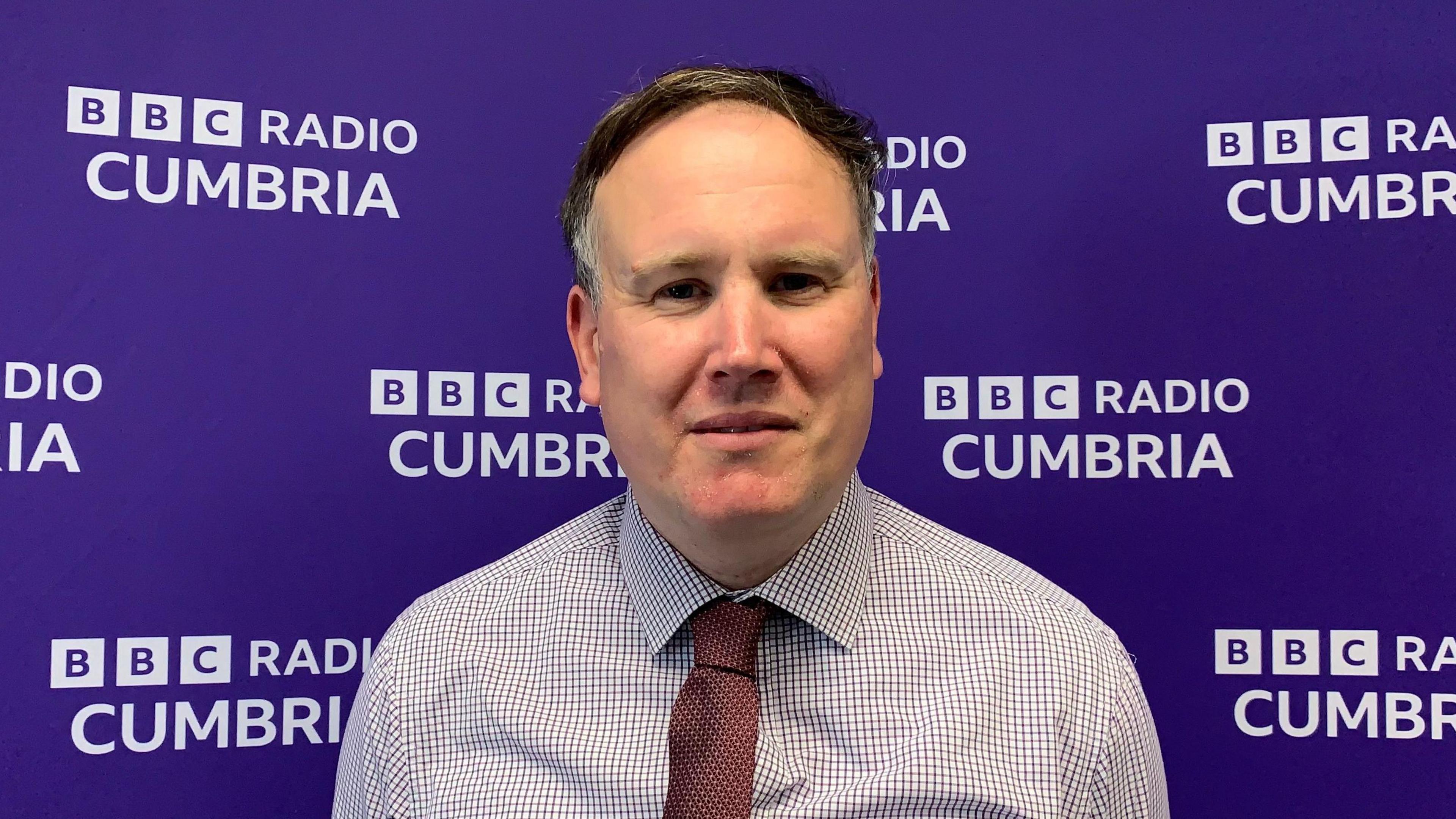 Owain Roberts has short grey/brown hair and is wearing a shirt and maroon tie. He is standing in front of a purple BBC Radio Cumbria branded backdrop.