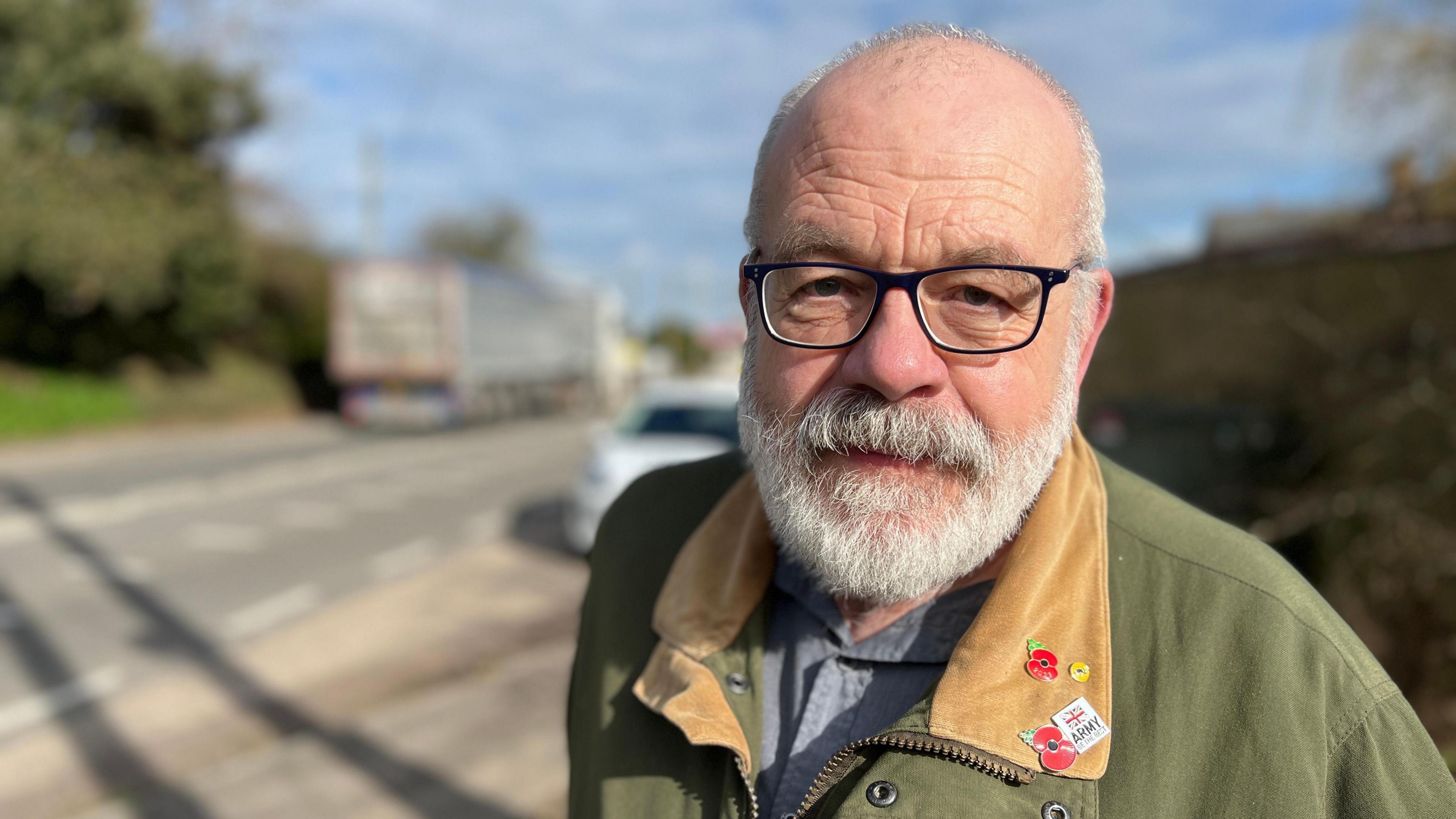 Andy Shuttleworth looks into the camera, bald with a grey beard and in front of the busy road