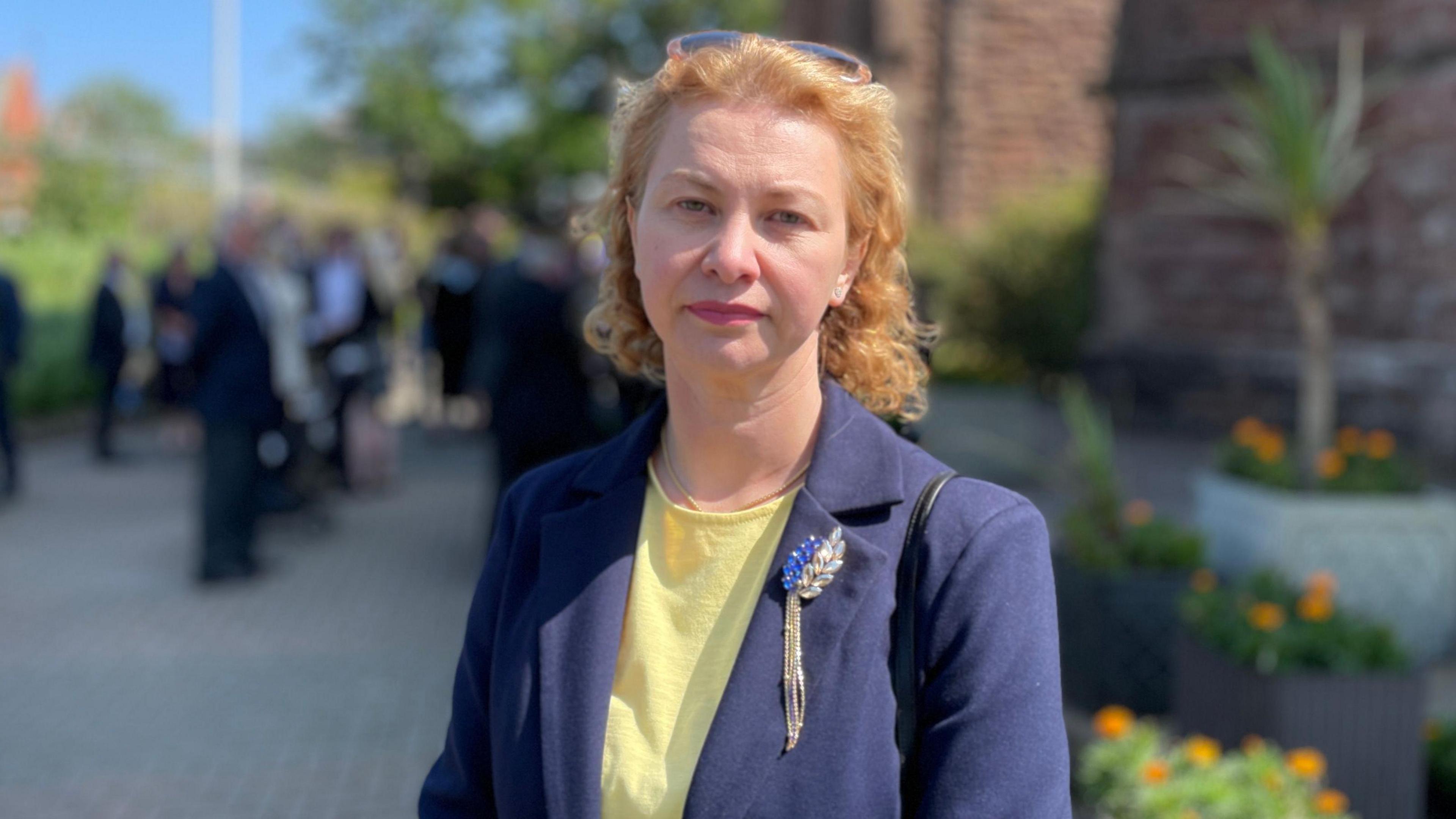 Karolina Davison, who has wavy shoulder length light coloured hair and is wearing a blue jacket over a yellow top. She is standing in the grounds of the cathedral.
