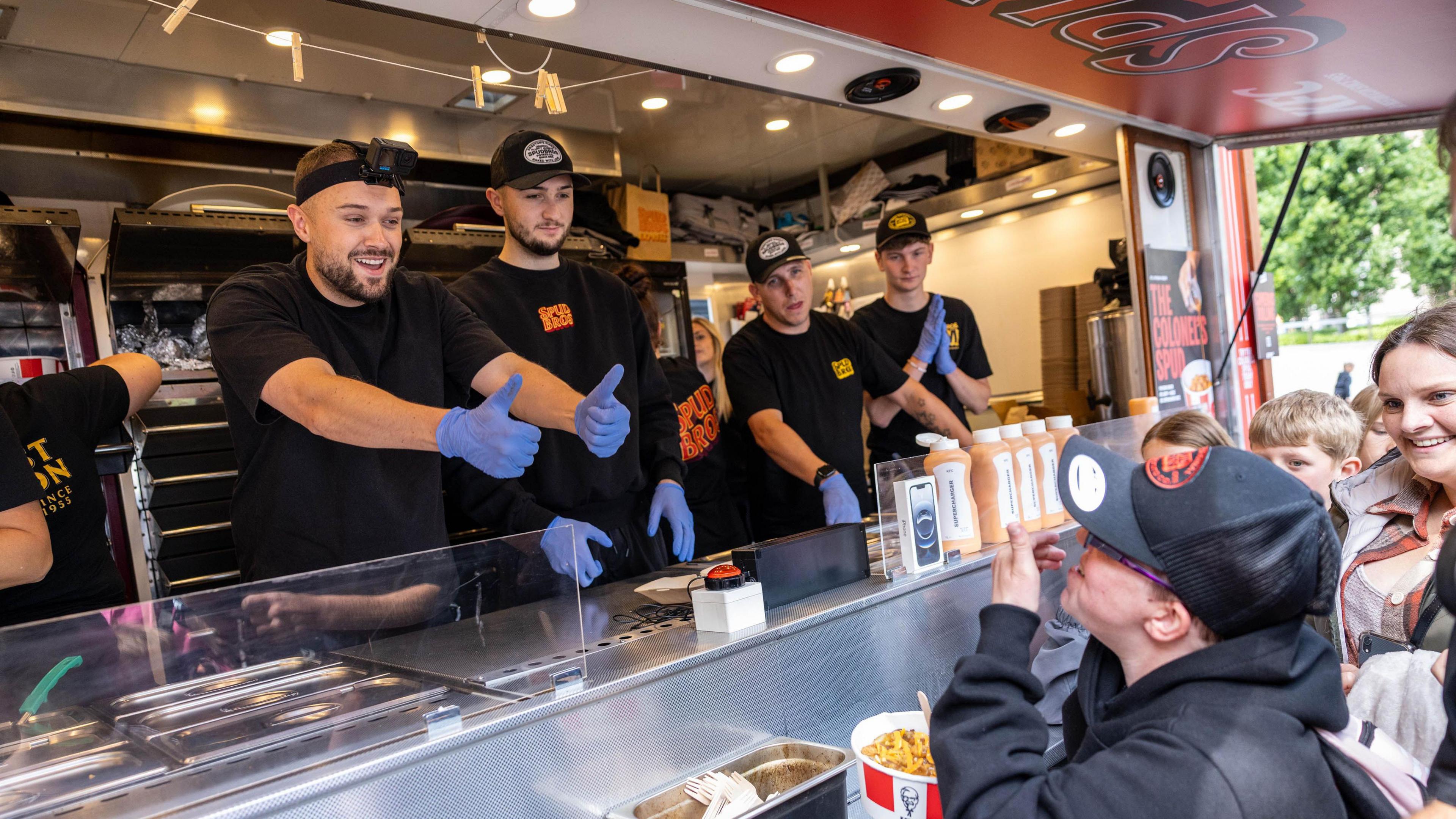 Roz Eccles holds his thumbs up from inside the serving hatch of the KFC catering van as a smiling child munches through some chicken at the front of the queue.