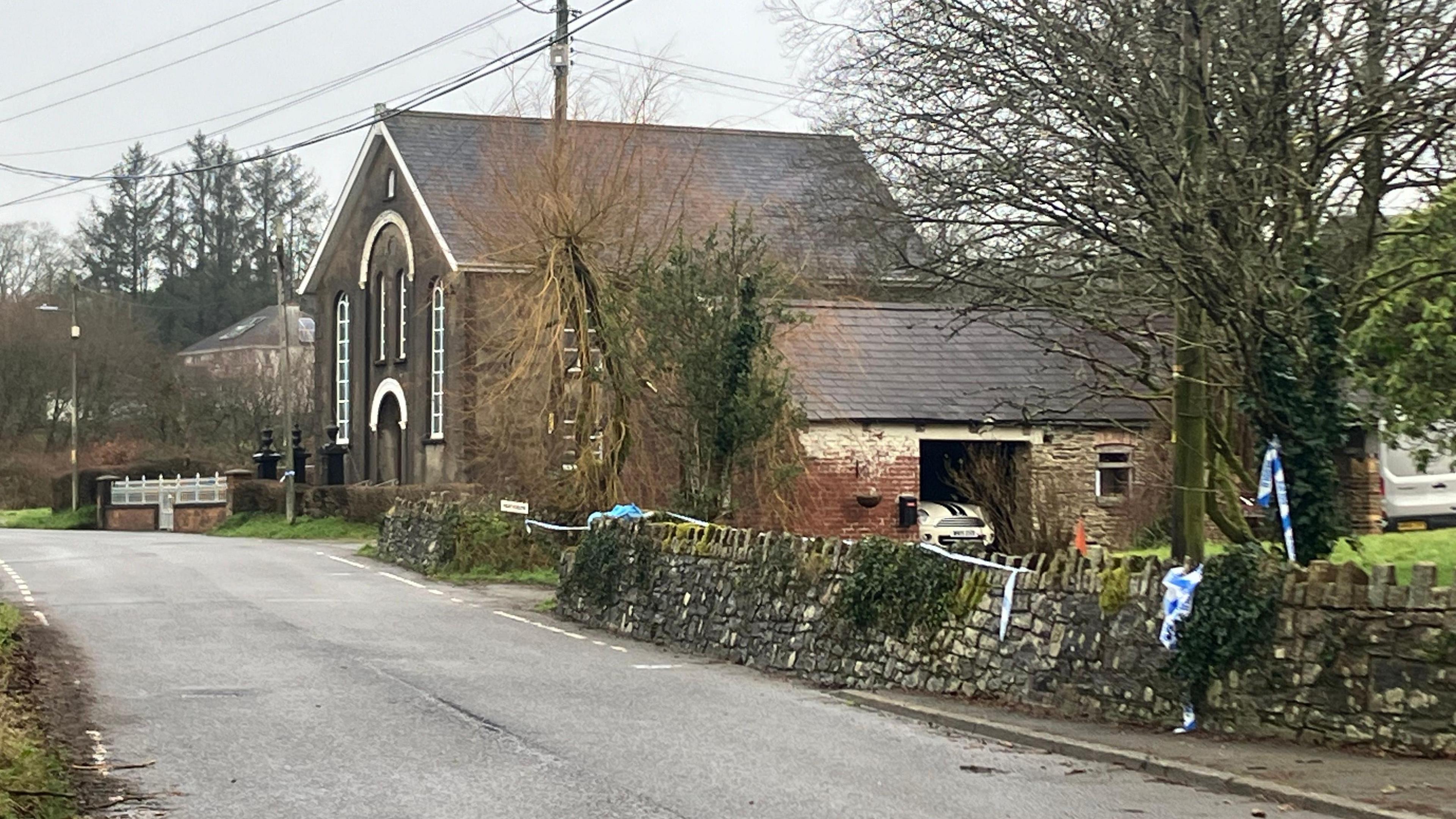 Road near Caer Salem Baptist Chapel. The chapel is visible and trees are in the background. Police tape is also visible across a driveway. 