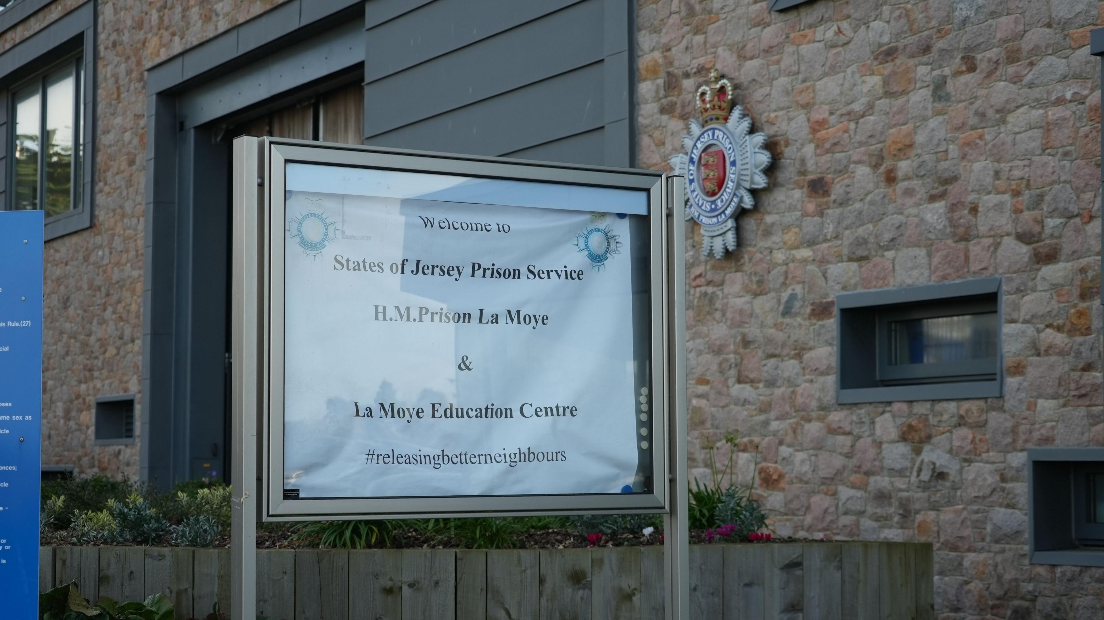 A sign outside the prison says States of Jersey Prison Service H.M. Prison La Moye and La Moye Education Centre with. Behind is the prison itself with bricks on the outter wall with the prison service logo on the wall.