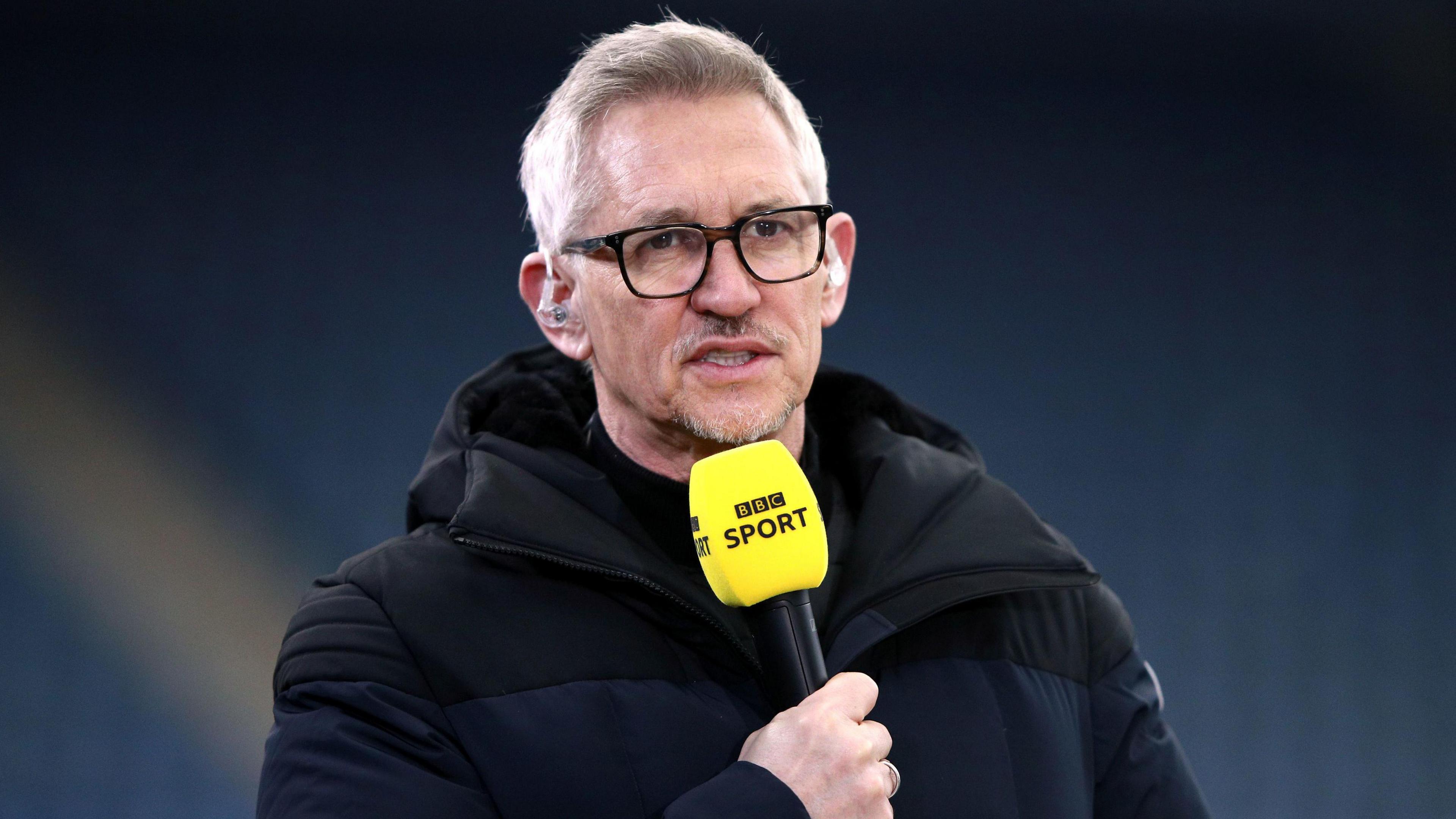 Gary Lineker, wearing a black coat, holds a BBC Sport microphone and wears headphones as he presents football coverage on TV 