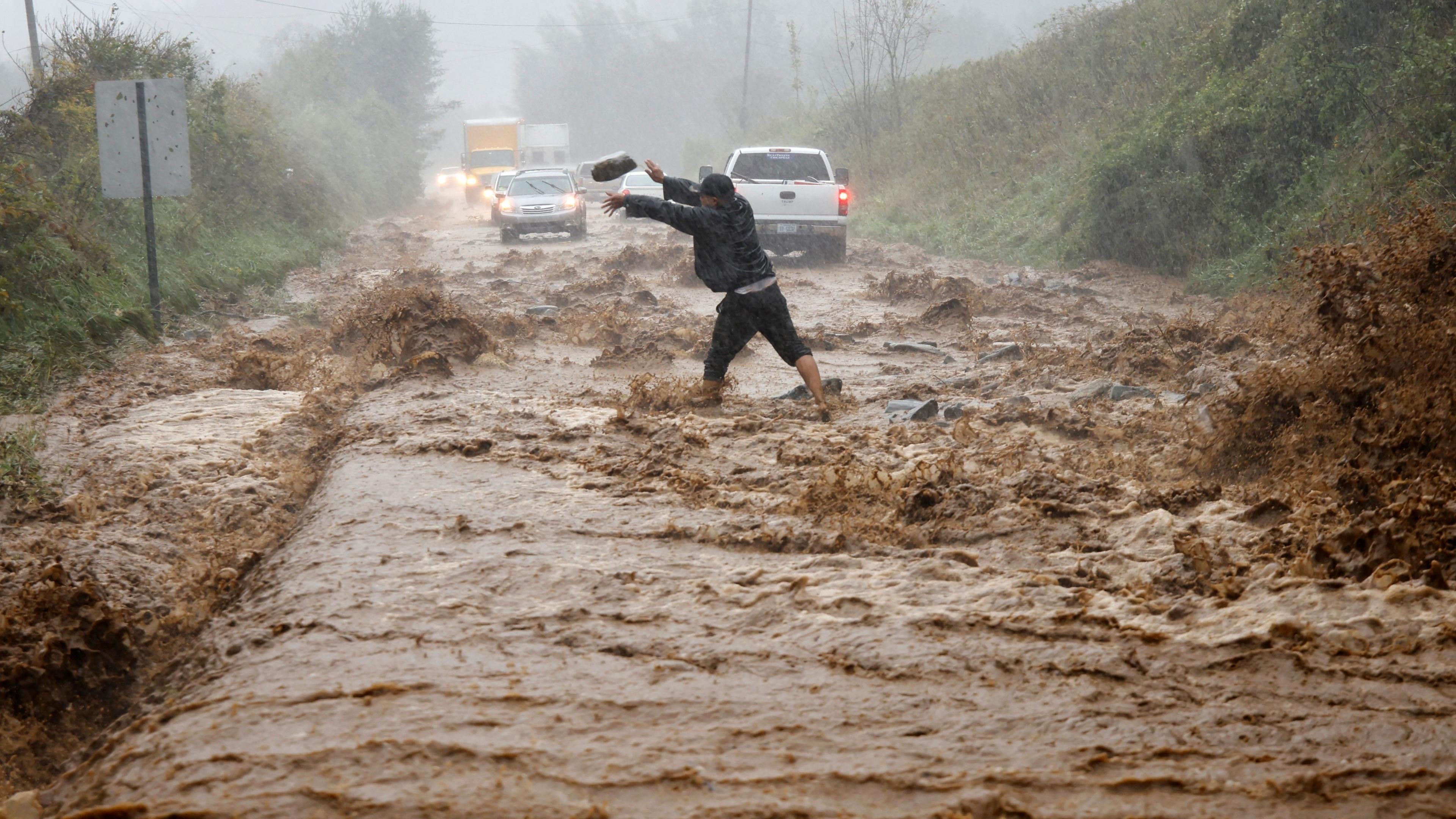 Man helps clear boulders from flooded road as cars negotiate floodwaters