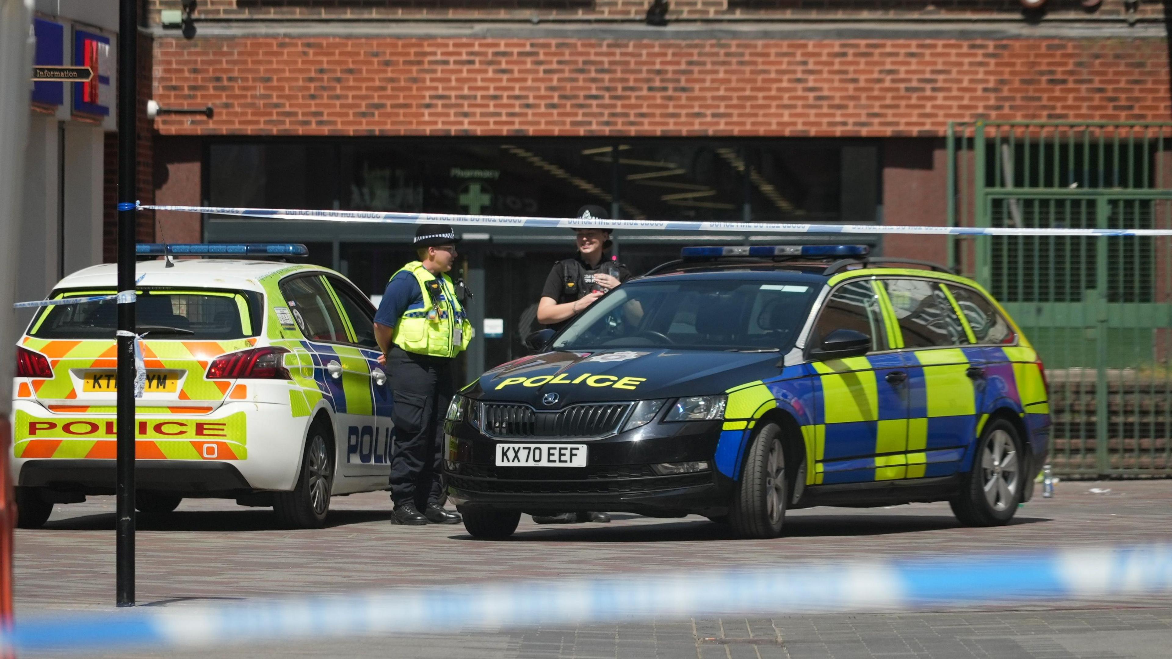 Two police cars are parked behind a blue and white police cordon. Two police officers stand between the cars talking to each other.
