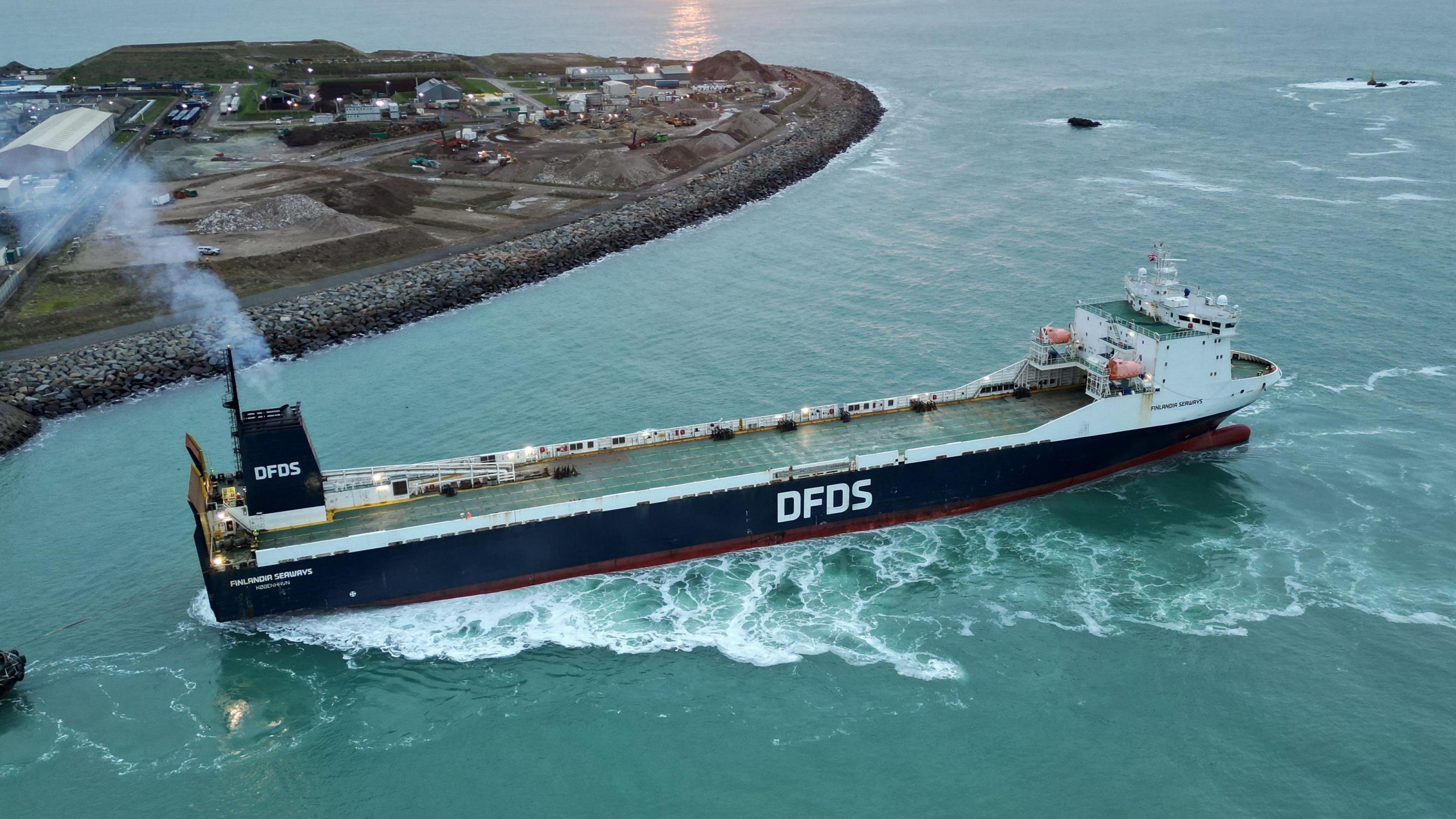 A long blue and white ferry with the letters DFDS on the side. A chimney is at the back of the ship with a light flow of smoke coming from the top.