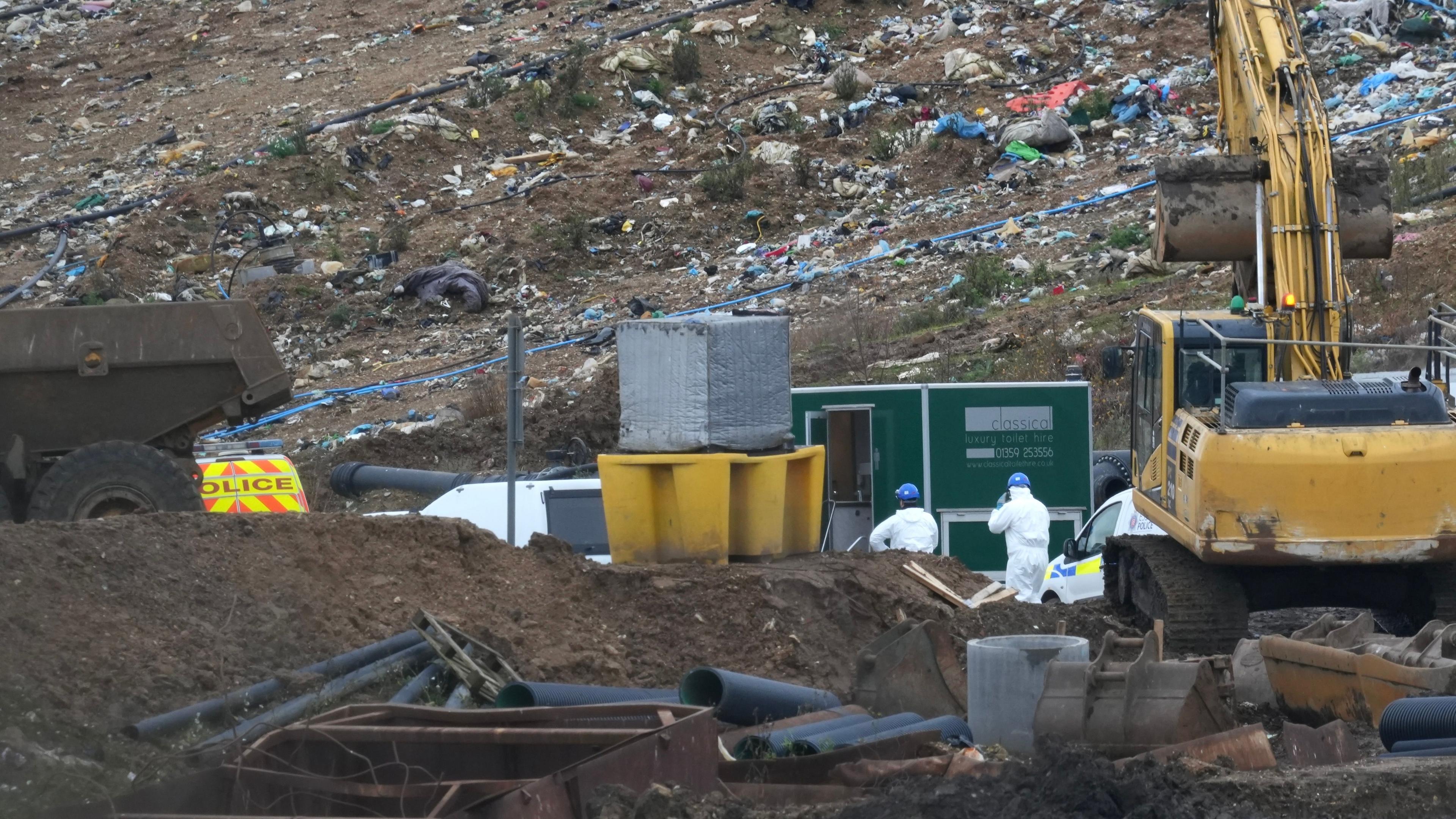 Two people in white body suits and blue hard hats in the middle of a landfill site, along with some police vans. Also pictured is rubbish, a digger and a portable toilet. 