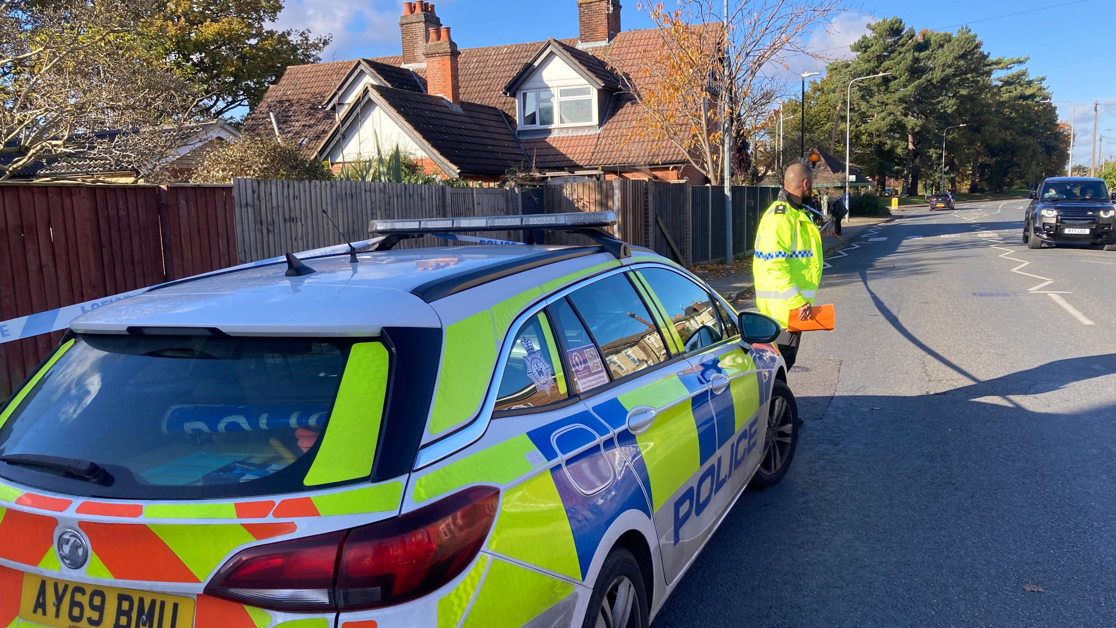 A road with a police car parked at the side of the road. There is police tape and an officer standing in front of the car in a yellow hi-vis police jacket